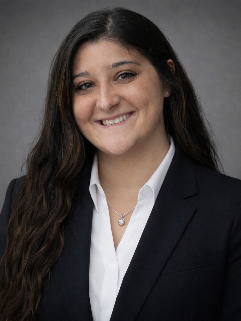 Professional portrait of a young woman with long brown hair, smiling, wearing a black blazer, white shirt, and a silver necklace with a pendant, against a gray background.