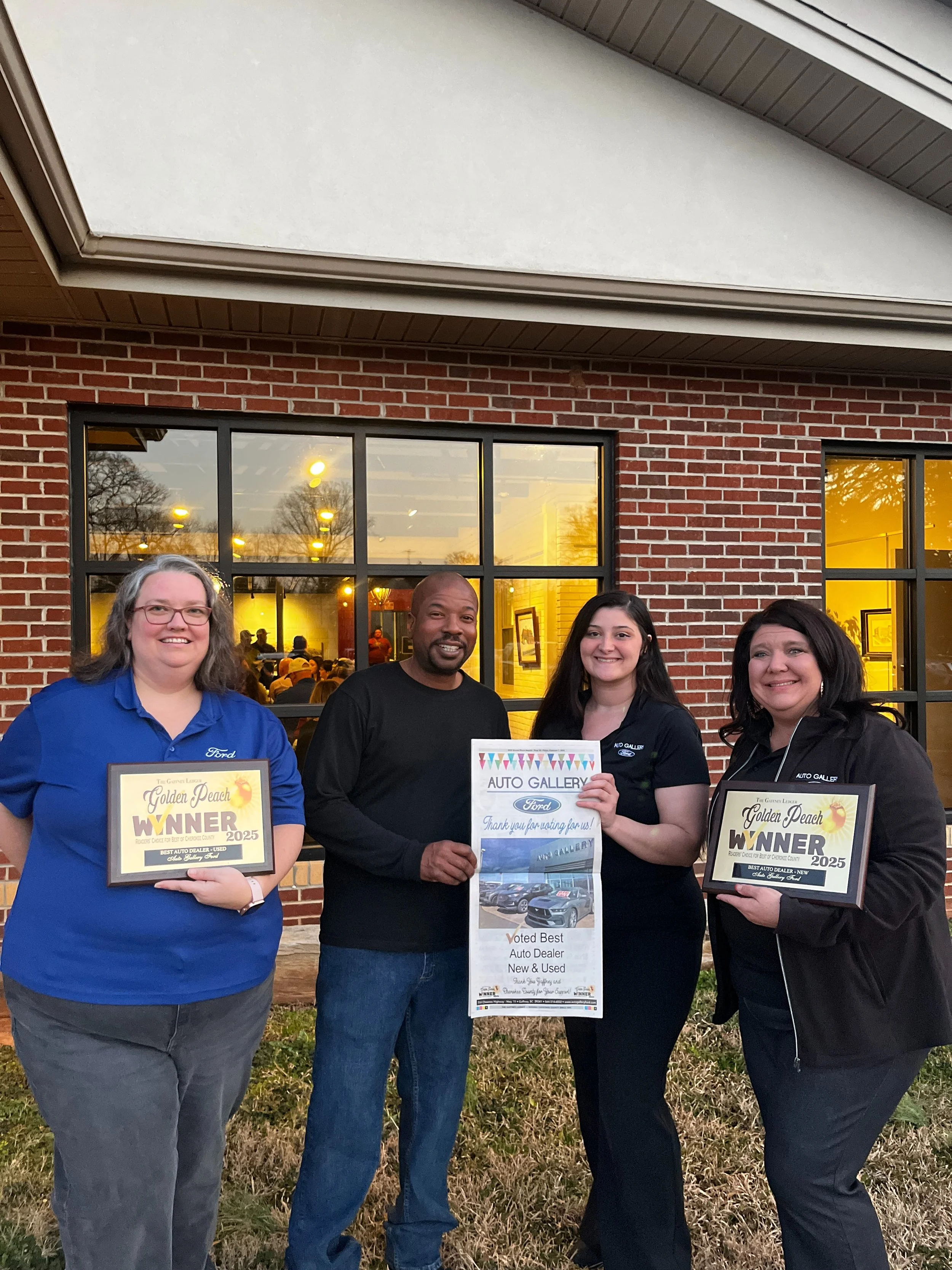 Four people standing outside in front of a brick building, holding awards and signs, smiling for a photo during an award ceremony or event.