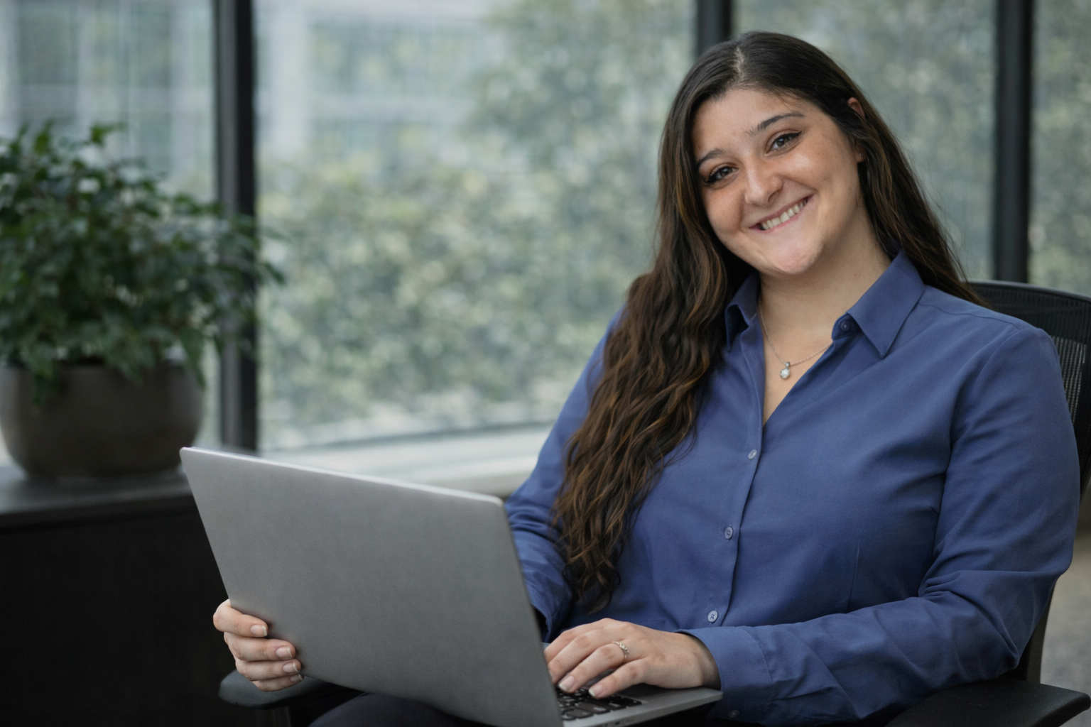 A young woman with long dark hair wearing a blue blouse, smiling while sitting at a desk and holding a laptop in front of a large window with a potted plant nearby.