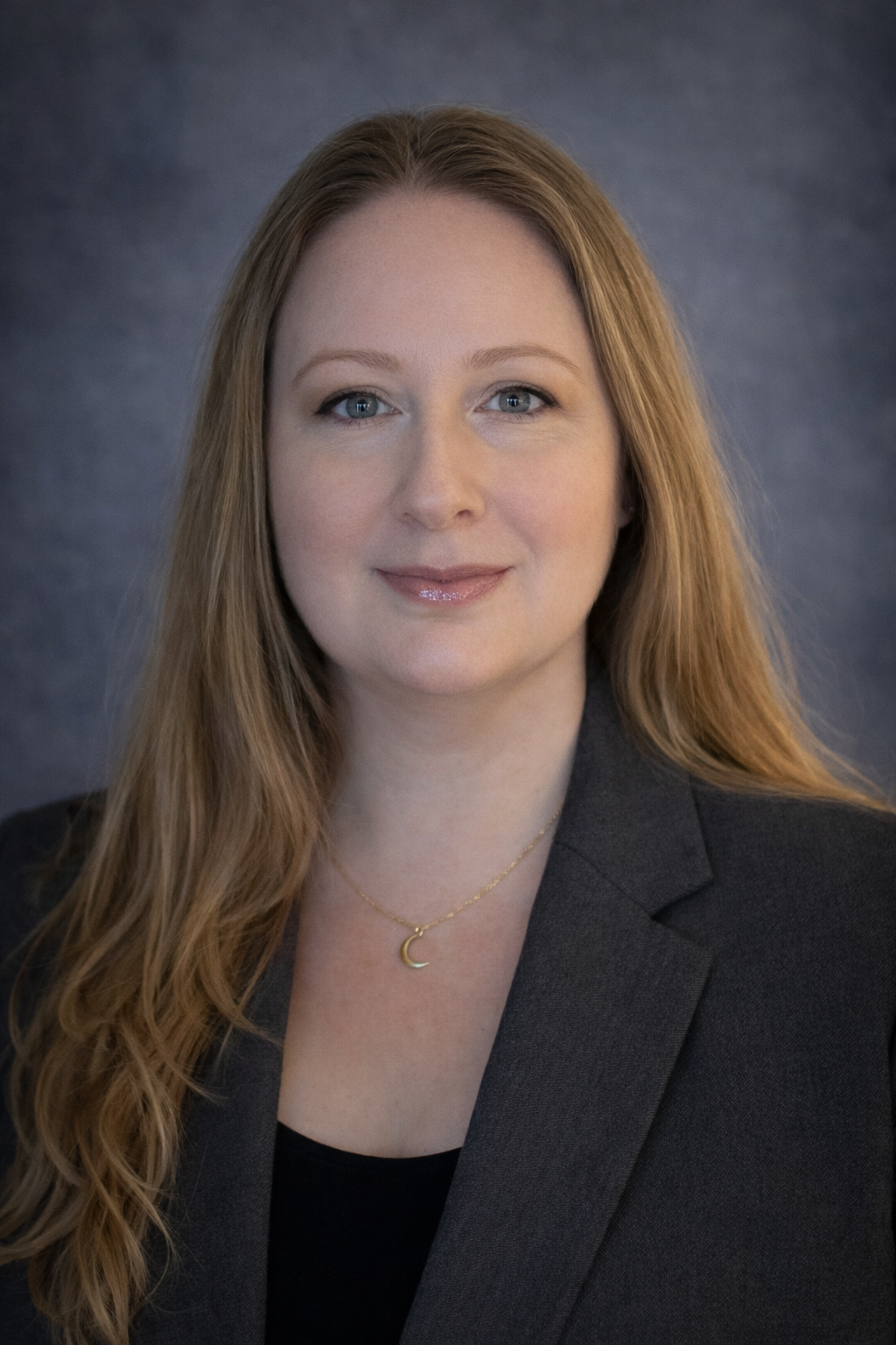 A woman with long red hair and blue eyes, wearing a black top and a dark blazer, smiling softly, with a gold crescent moon necklace, standing against a dark gray background.