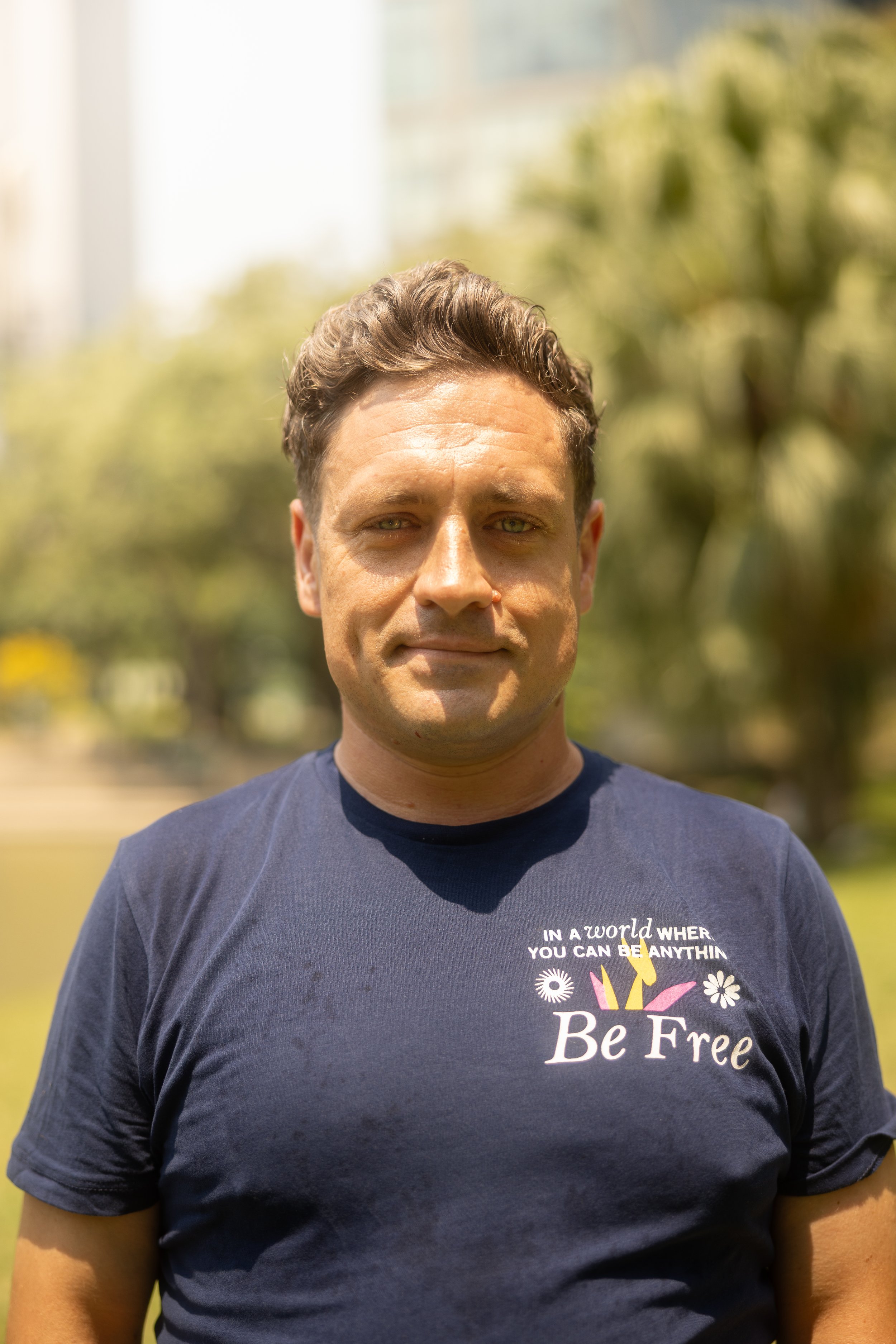 A man standing outdoors with a blurred background of trees. He is wearing a dark blue t-shirt with the words "Be Free" and a colorful design on it. The man has short, styled brown hair and green eyes.