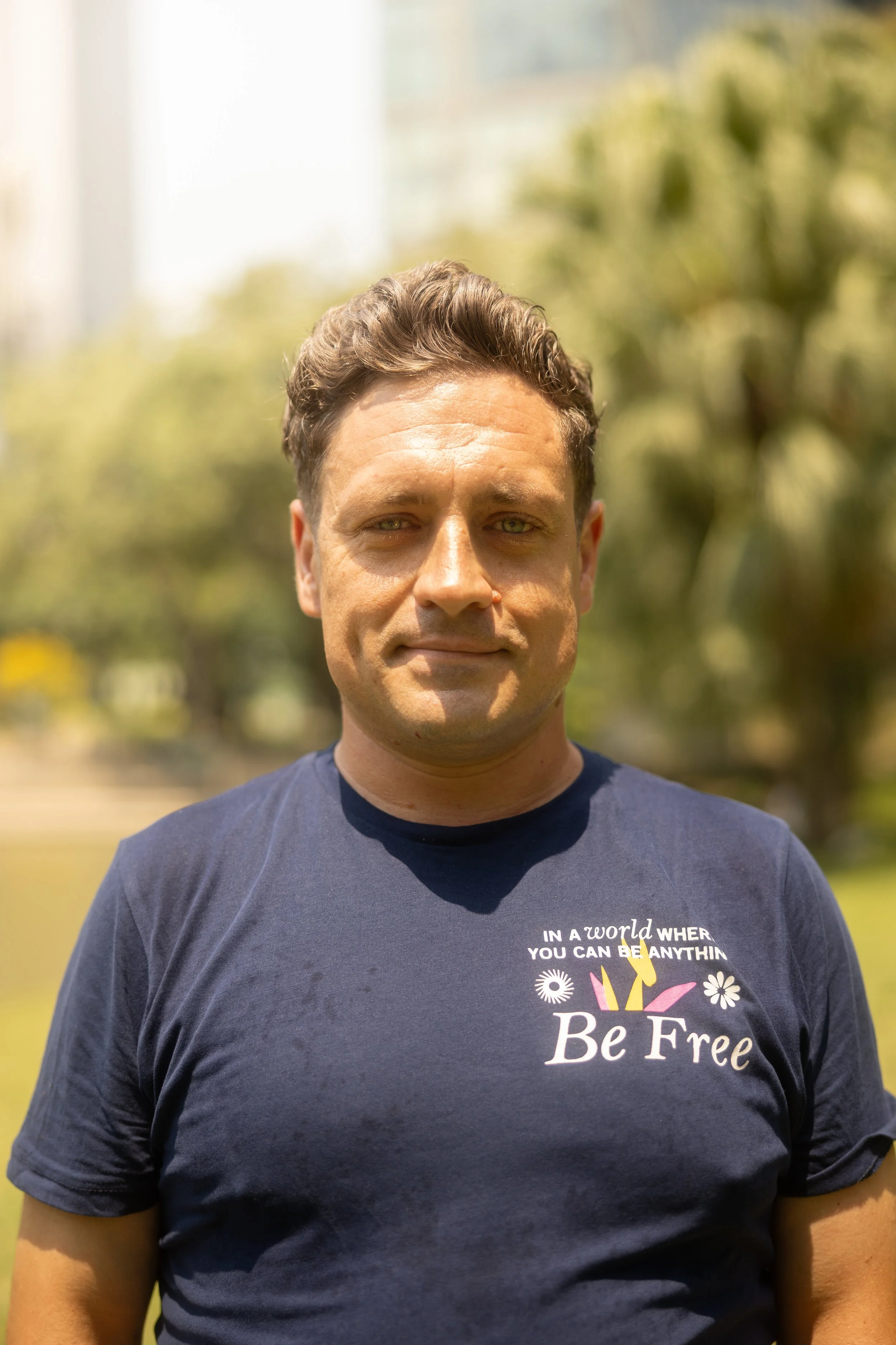 A man with light brown hair and green eyes is standing outdoors in a park or garden, wearing a navy blue T-shirt with a message that says 'In a world where you can be anything, Be Free.' The background is blurred with greenery and sunlight.