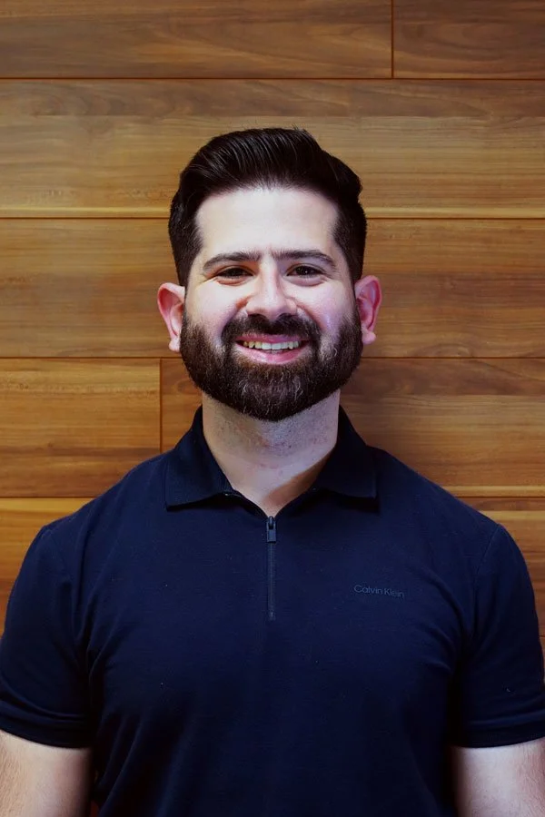 A smiling man with dark hair and a beard, wearing a navy blue Calvin Klein polo shirt, standing in front of a wooden wall.