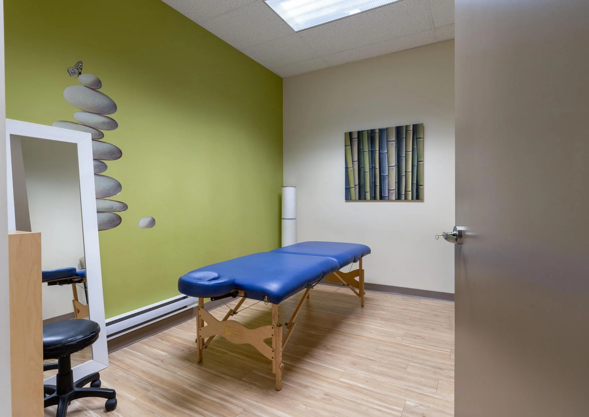 Medical examination room with a massage or examination table, a chair, a floor-length mirror, and wall art of stacked stones and bamboo, with a green accent wall and wood flooring.