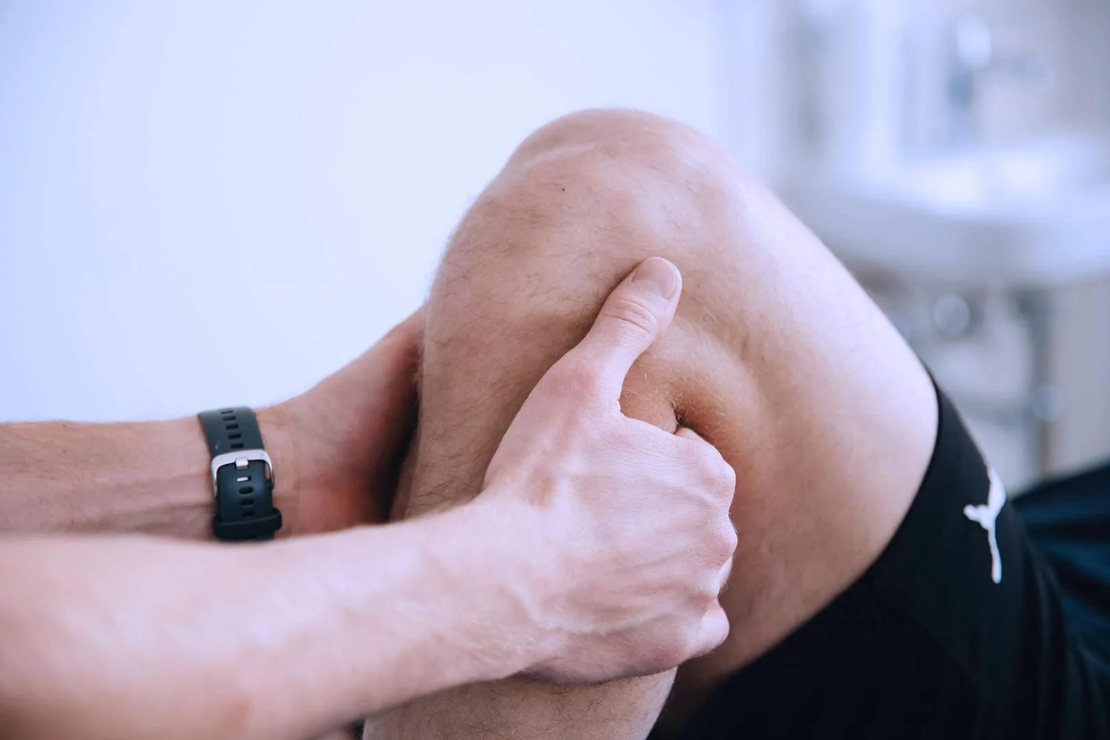 A person is holding and examining their knee, which is bent. The person is wearing a black watch on their left wrist and black shorts.