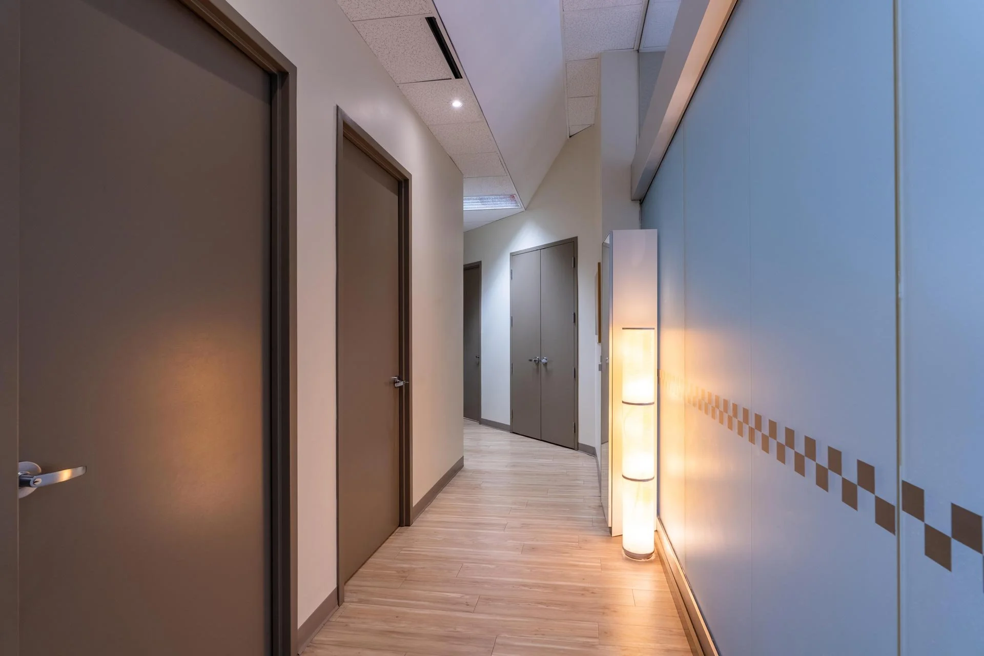 A hallway with beige walls, brown doors, light wood flooring, and modern lighting, including a tall, cylindrical floor lamp.