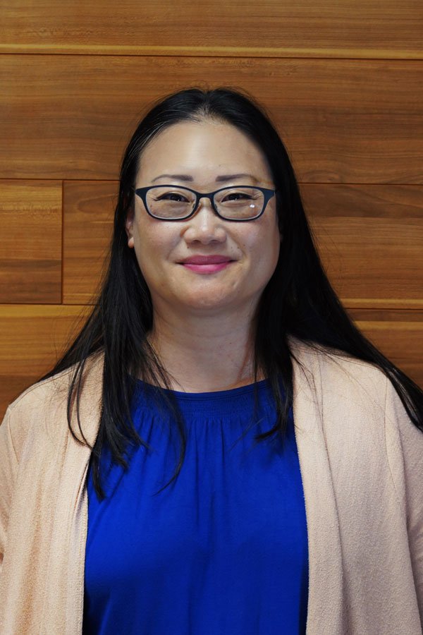 A woman with long black hair, wearing glasses, a royal blue blouse, and a beige cardigan, standing in front of a wooden wall.