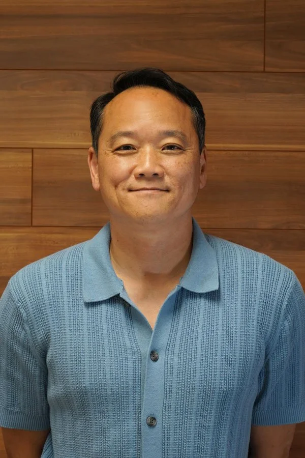 Man in a blue short-sleeve polo shirt smiling in front of a wooden wall.