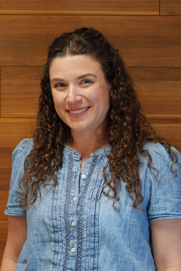 A woman with long curly brown hair smiling, wearing a light blue denim shirt, with a wooden wall background.