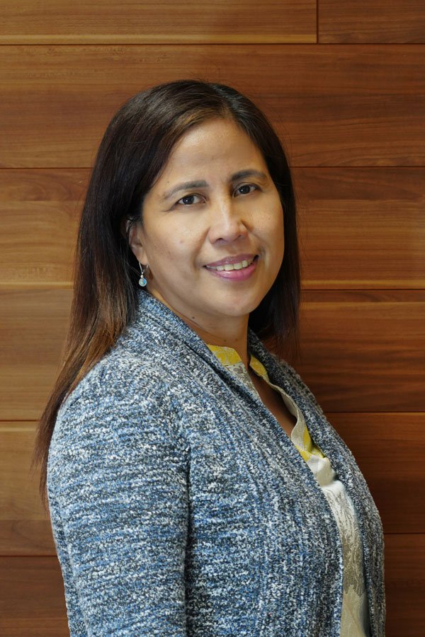 A woman with long dark hair smiling, wearing a gray blazer and a patterned yellow and white blouse, standing in front of a wooden wall.