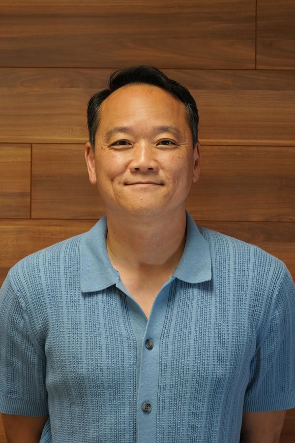A smiling man with short black hair wearing a light blue knit polo shirt against a wooden wall background.