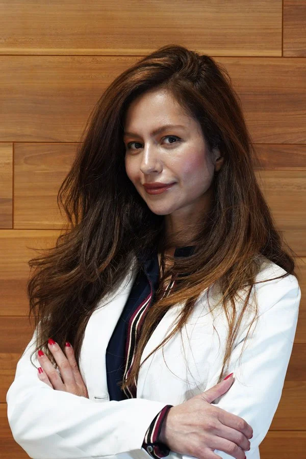 A woman with long brown hair and fair skin standing in front of a wooden wall, wearing a white blazer and a dark top, with her arms crossed and a slight smile.