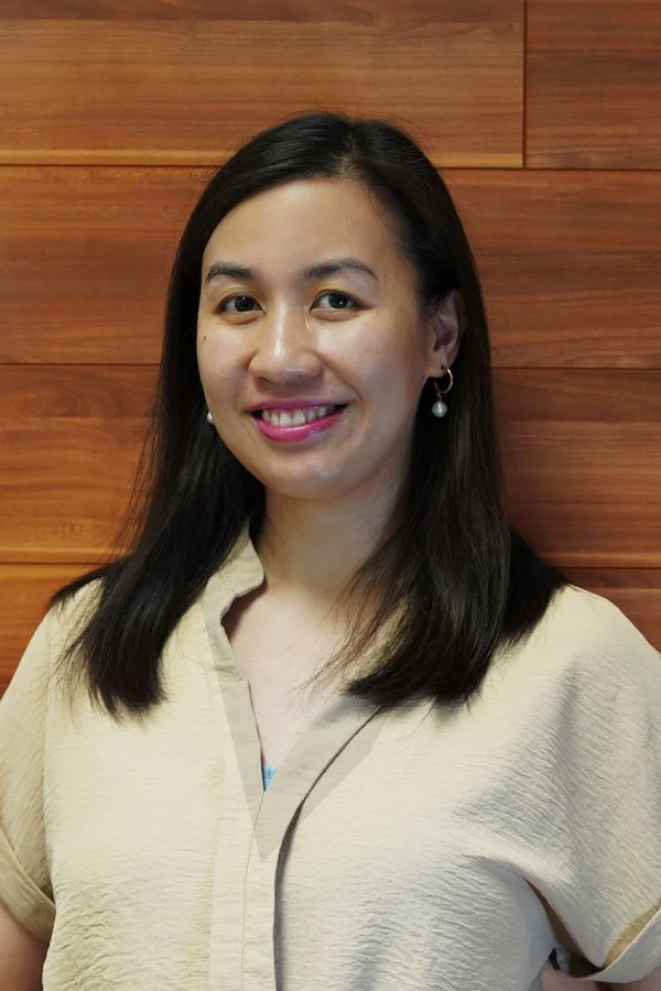 A young woman with long dark hair, smiling, wearing earrings and a beige blouse, standing in front of a wooden wall.