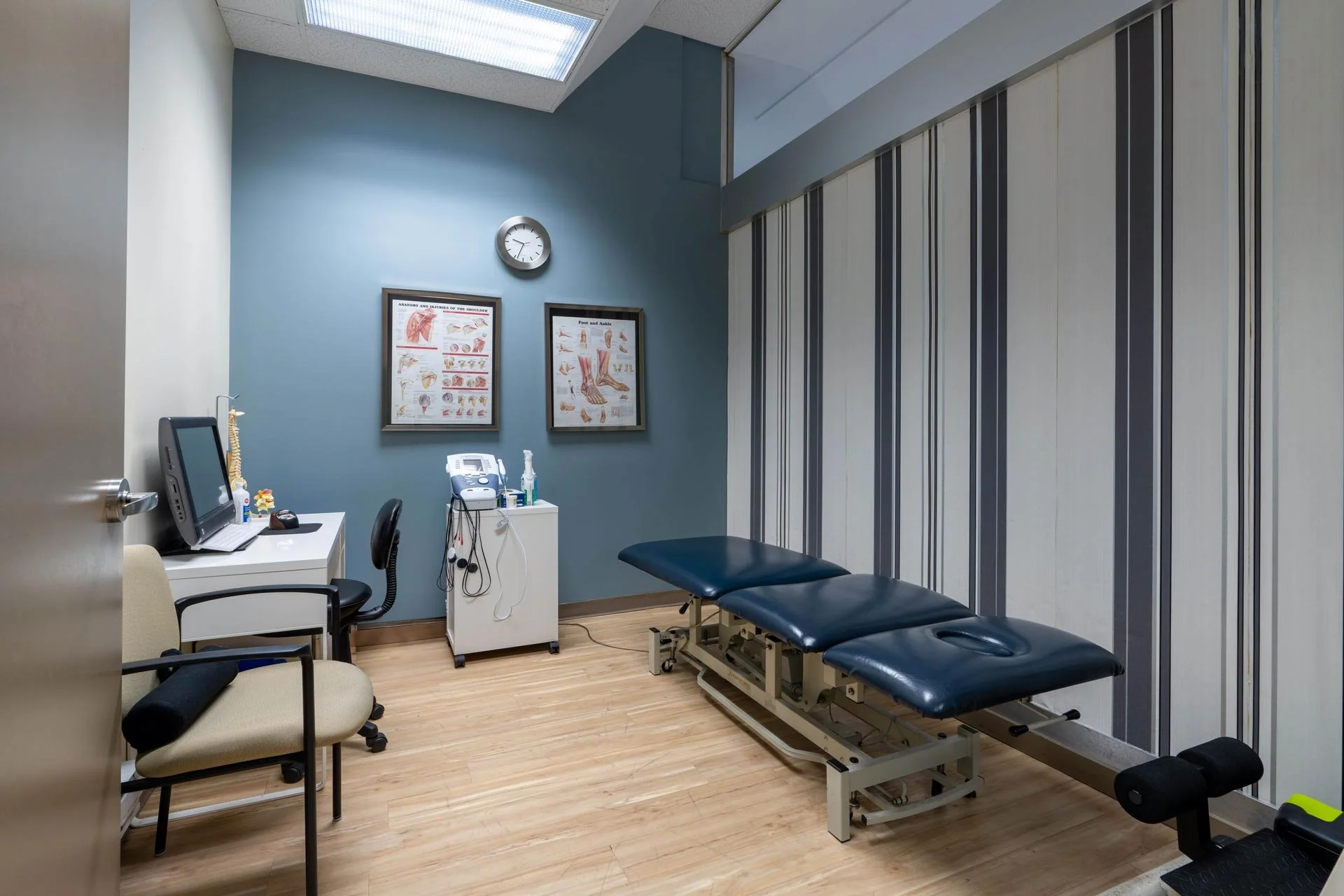 Medical examination room with a patient table, chairs, computer, medical charts on the wall, and medical equipment, illuminated by overhead fluorescent lighting.