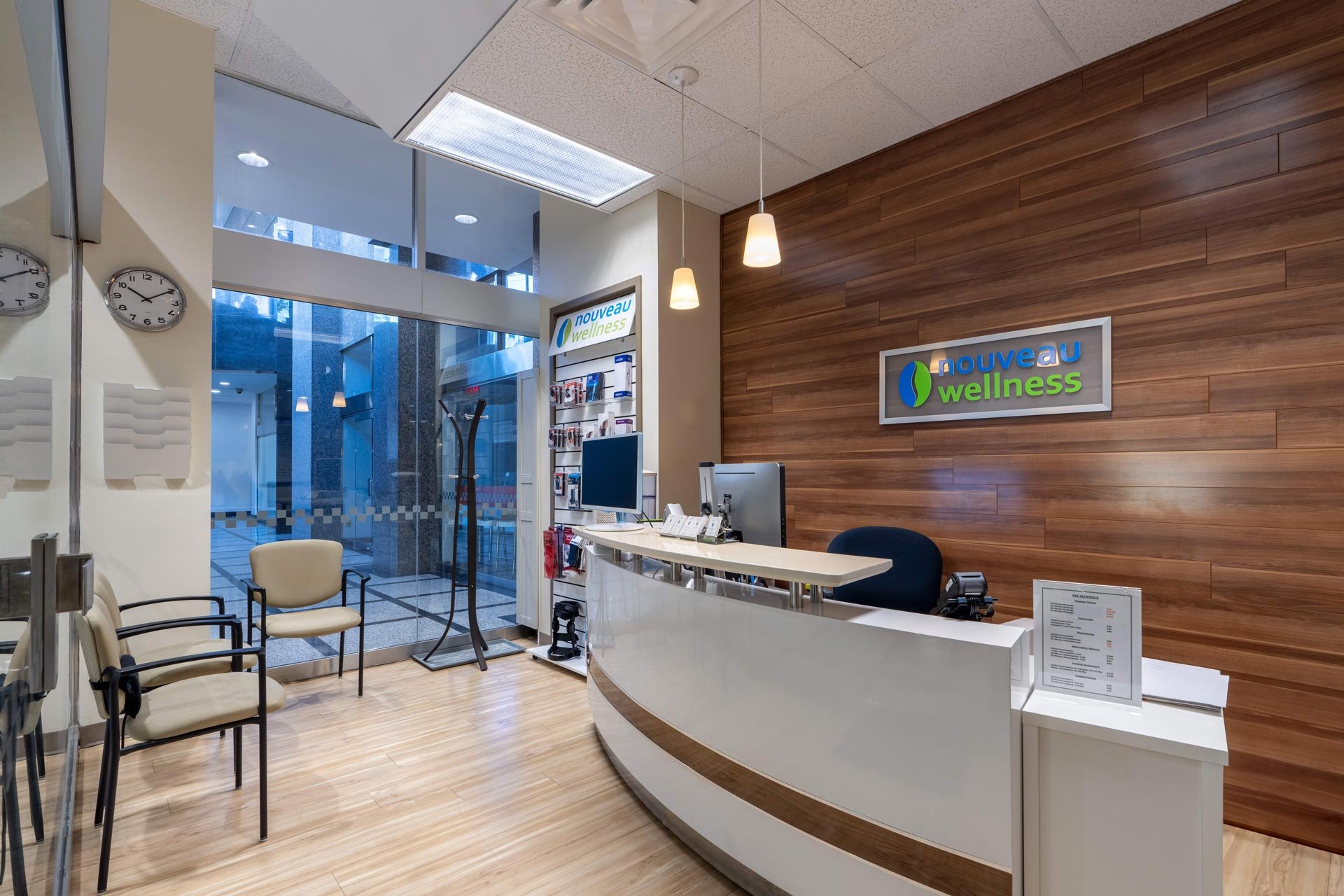 Reception area of a wellness or medical clinic with a wooden accent wall, sitting chairs, a curved white reception desk, and a sign that reads 'Nouveau Wellness'.