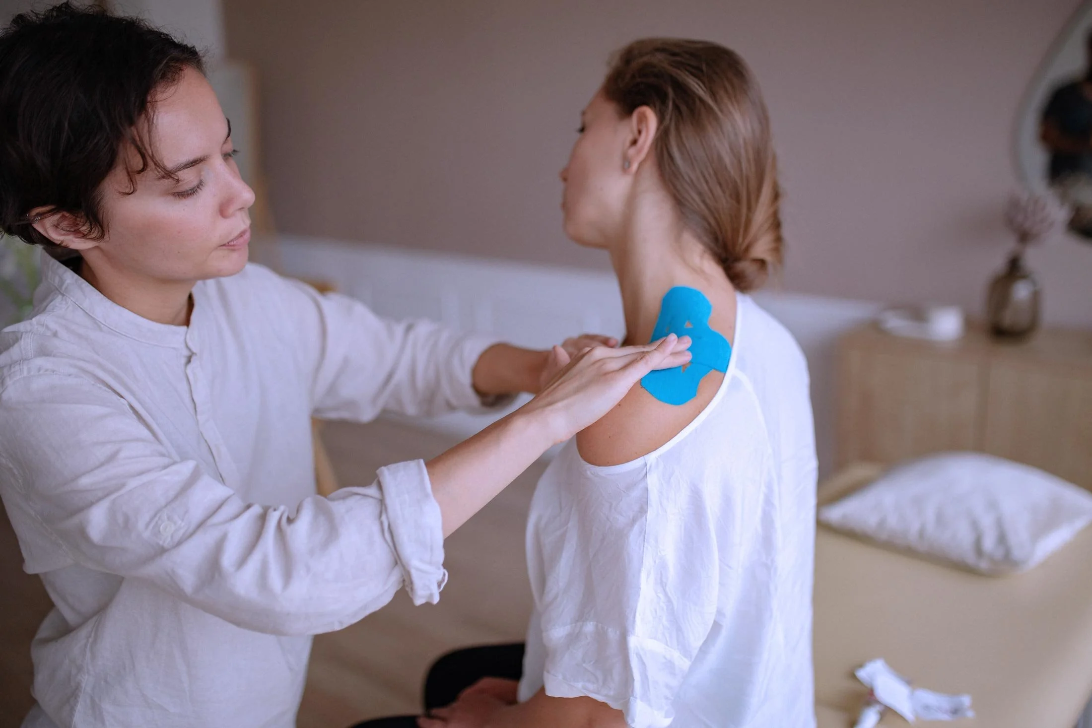 A woman with short dark hair and wearing a white shirt is applying blue kinesiology tape to the shoulder of a young girl with reddish hair in a ponytail, in a room with a neutral wall and a lamp in the background.