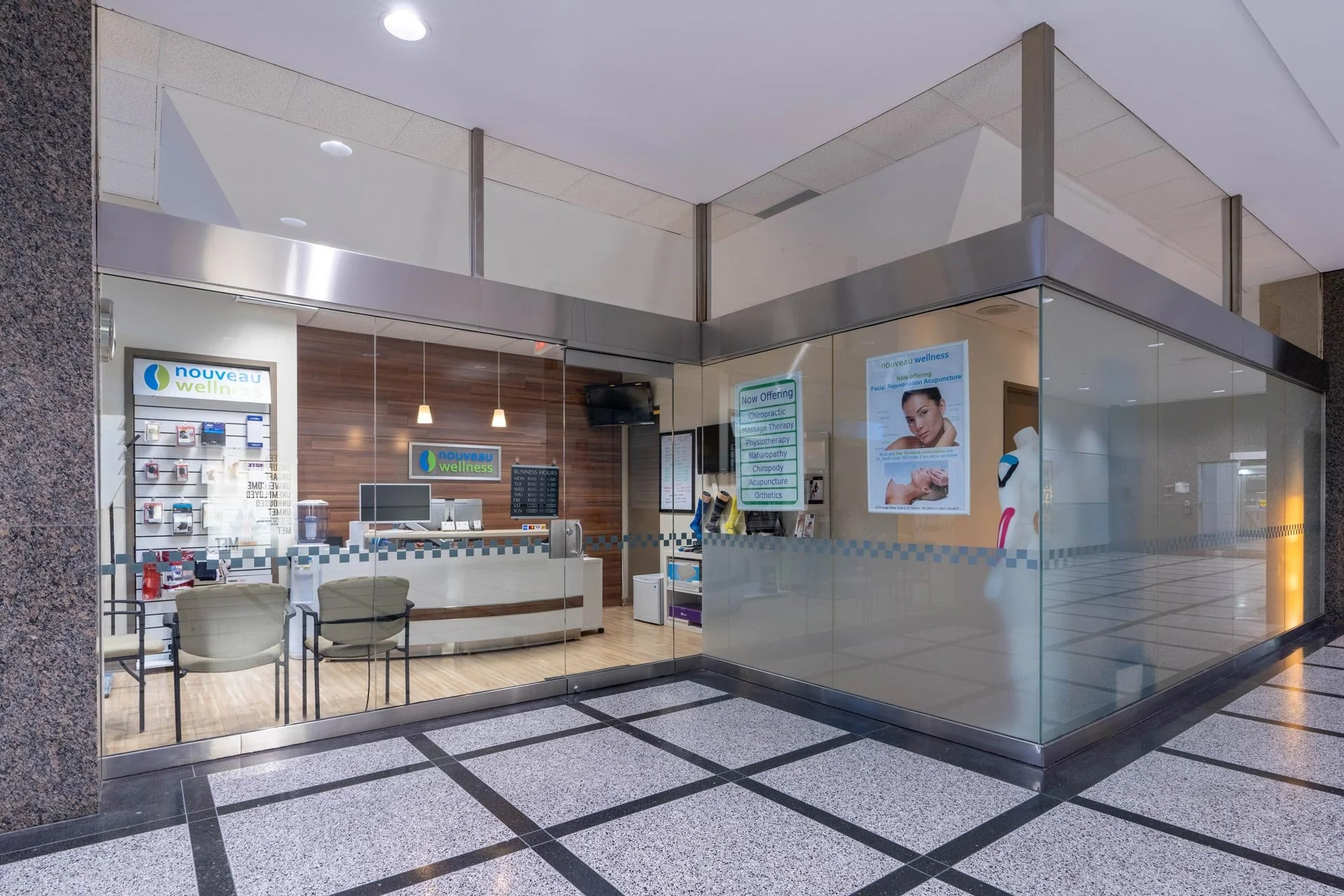 Exterior view of a wellness clinic inside a shopping mall, featuring glass walls, waiting area with chairs, and promotional signs for various health services like chiropractic and acupuncture.