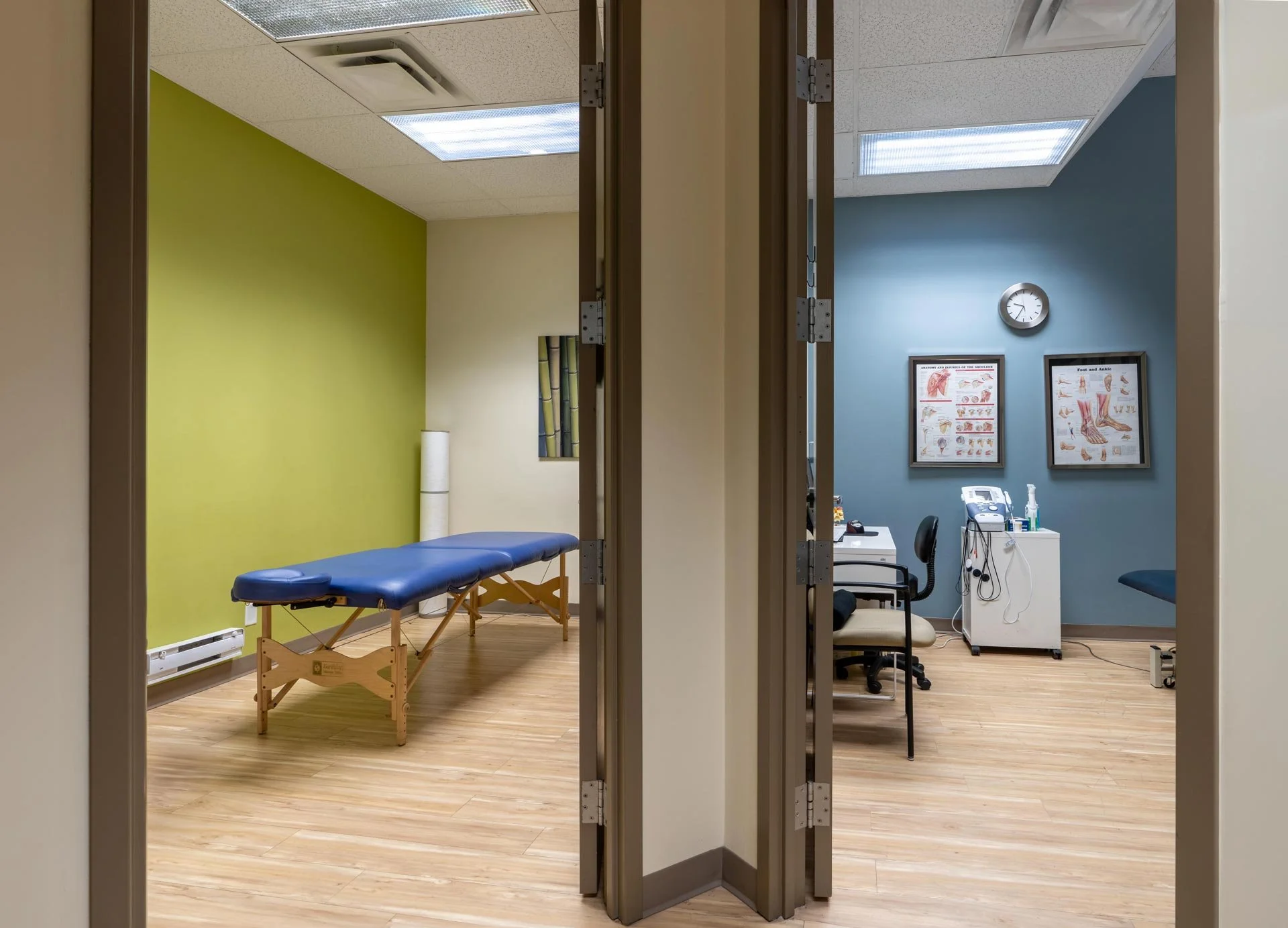 View of a medical examination room with a blue cushioned examination table against a lime-green wall, and a blue wall with anatomical and foot and ankle diagrams, clock, desk, chair, and medical equipment. The room has wooden flooring and is seen thr