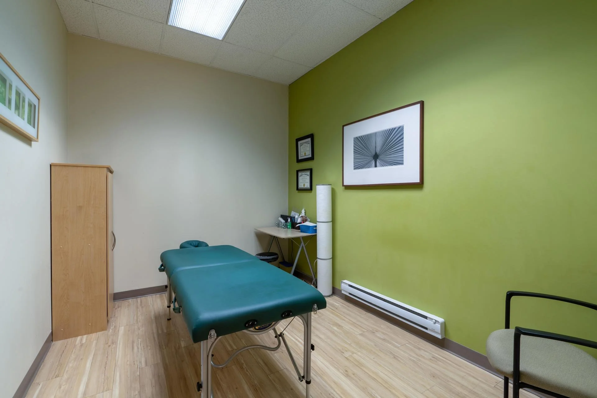 A medical examination room with a green wall on the right and a beige wall on the left. There is a green examination table in the center, a wooden cabinet on the left, and a small table with supplies on the right. The room has framed certificates and