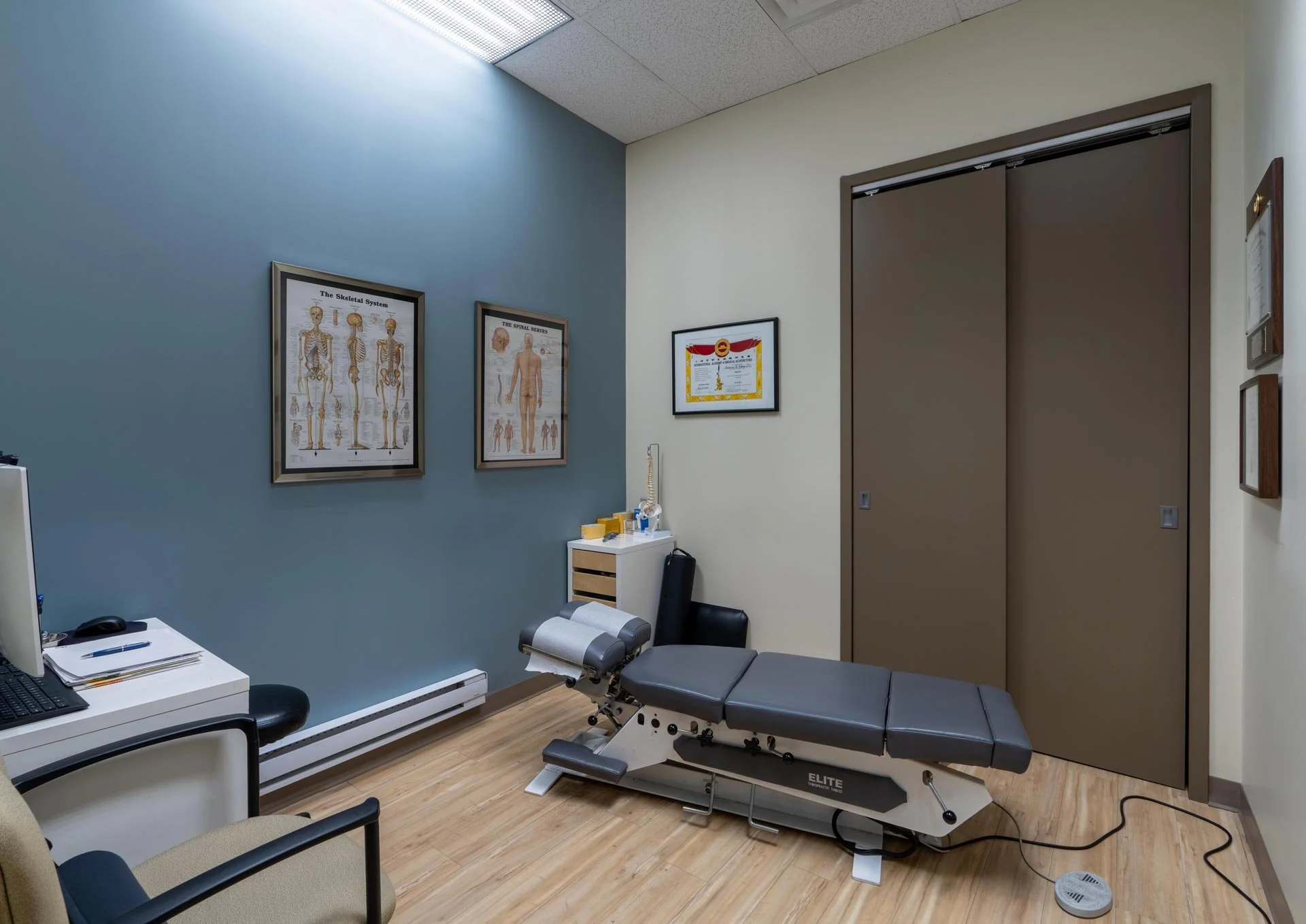Medical examination room with examination table, chairs, computer, and anatomical posters on the wall.