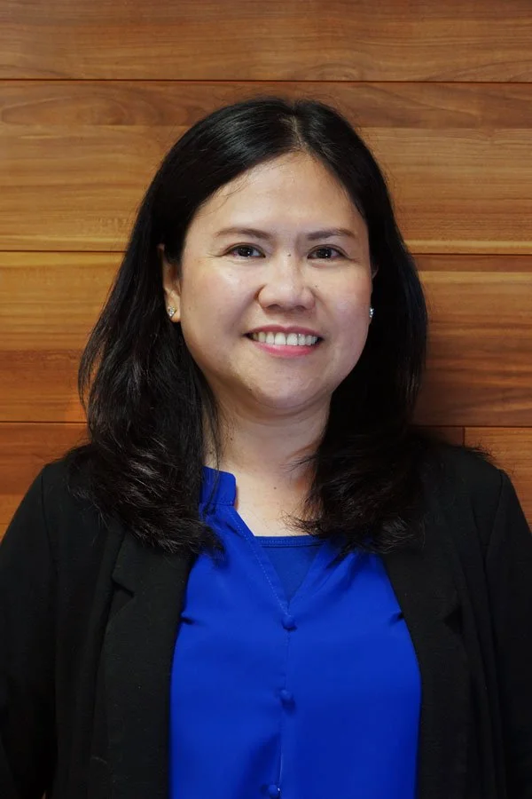 A woman with shoulder-length black hair smiling, wearing a blue blouse and black blazer, standing in front of a wooden wall.