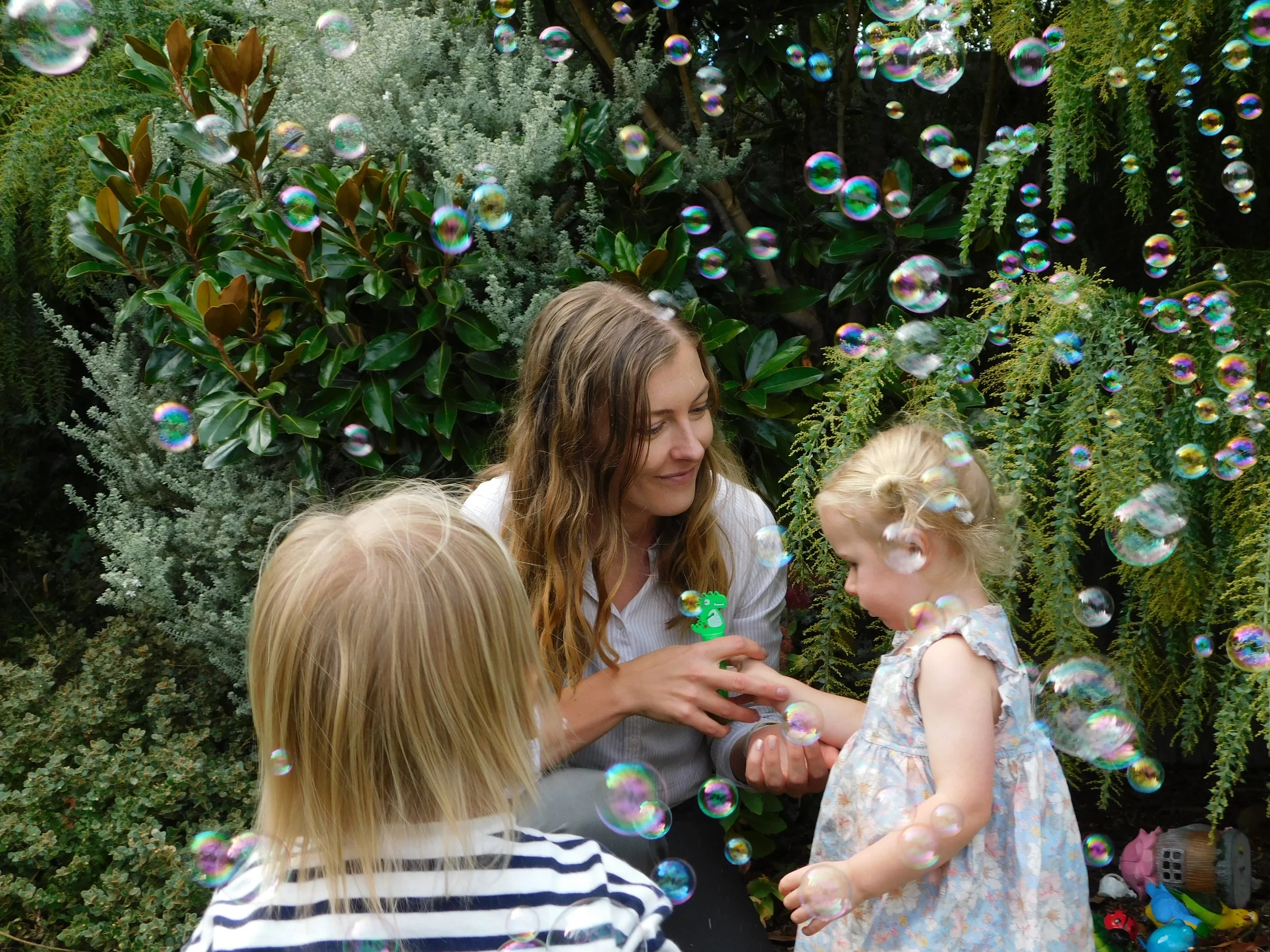 A woman with long curly hair and light skin holding a young girl with braided blonde hair and light skin, surrounded by children in a garden with bubbles floating around. The woman appears to be showing something to the girl while other kids look on.