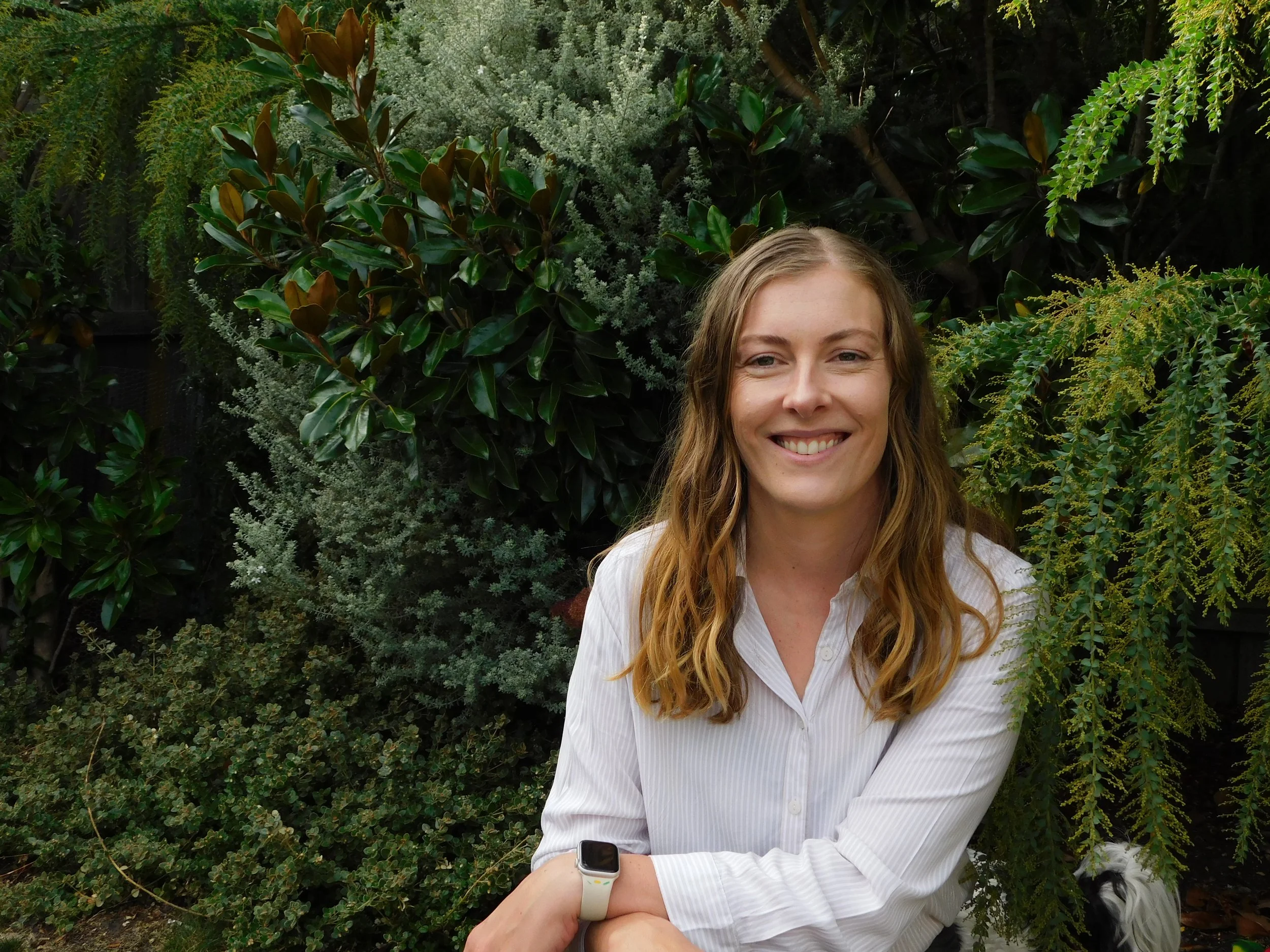 A woman with long, wavy red hair smiling in front of green bushes and trees, wearing a white button-up shirt and a smartwatch on her left wrist.
