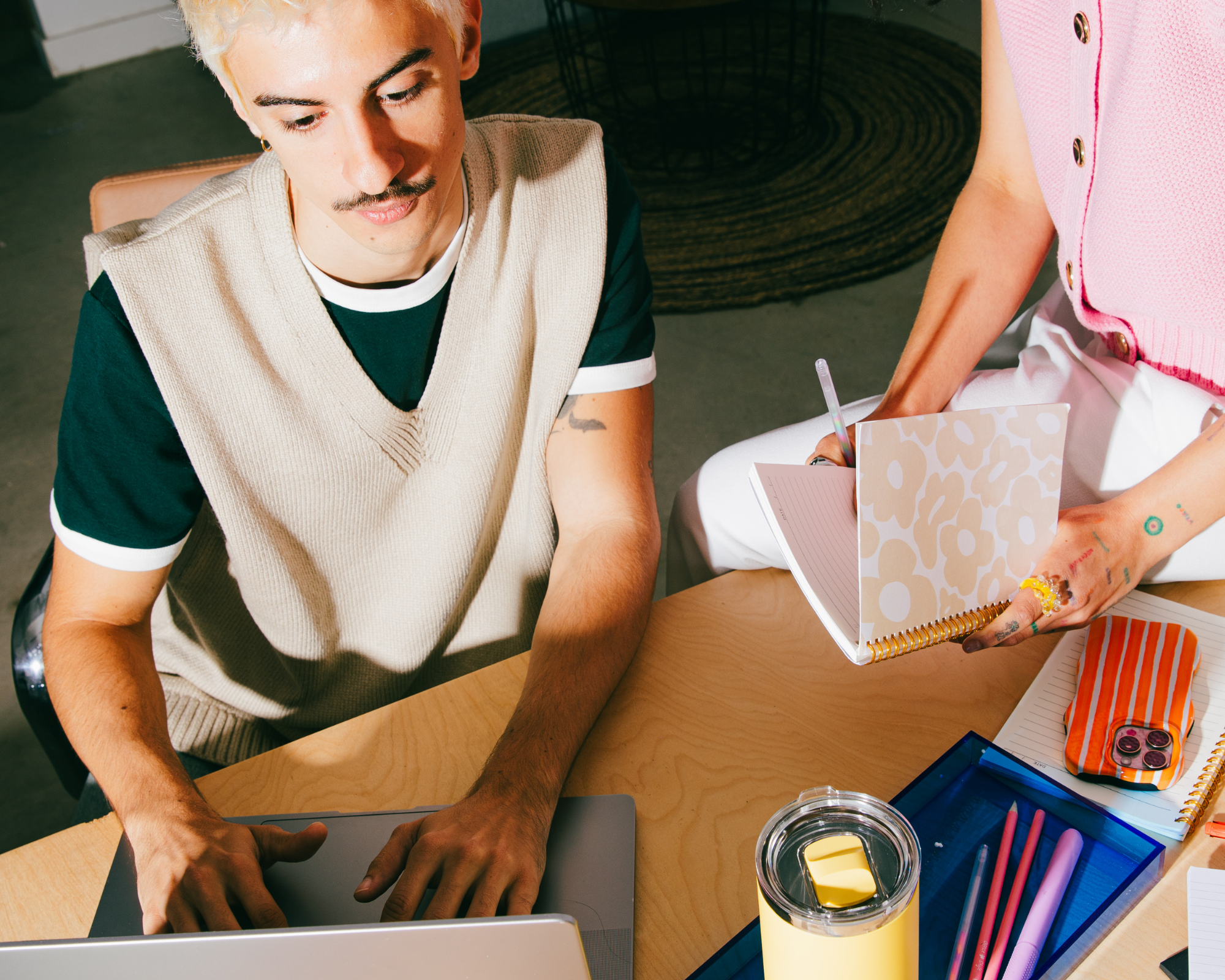 A man with platinum blonde hair and a mustache sitting at a wooden table with various stationery items, a closed laptop, and a person's hand reaching over.