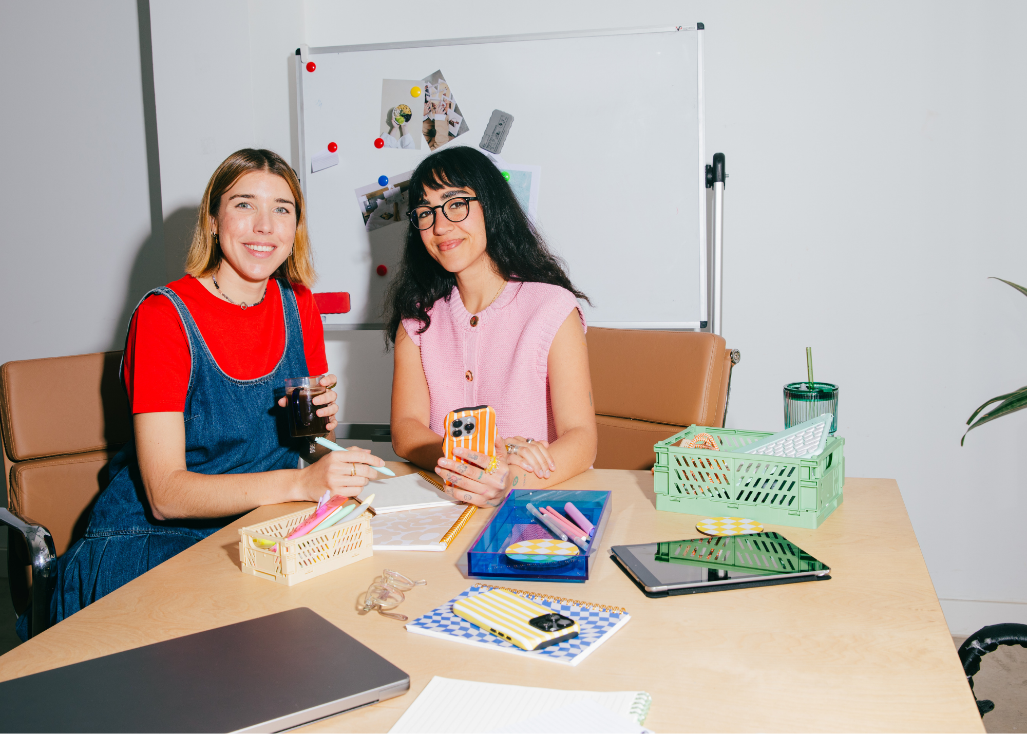Two women sitting at a table in a meeting room, surrounded by colorful office supplies and a whiteboard.