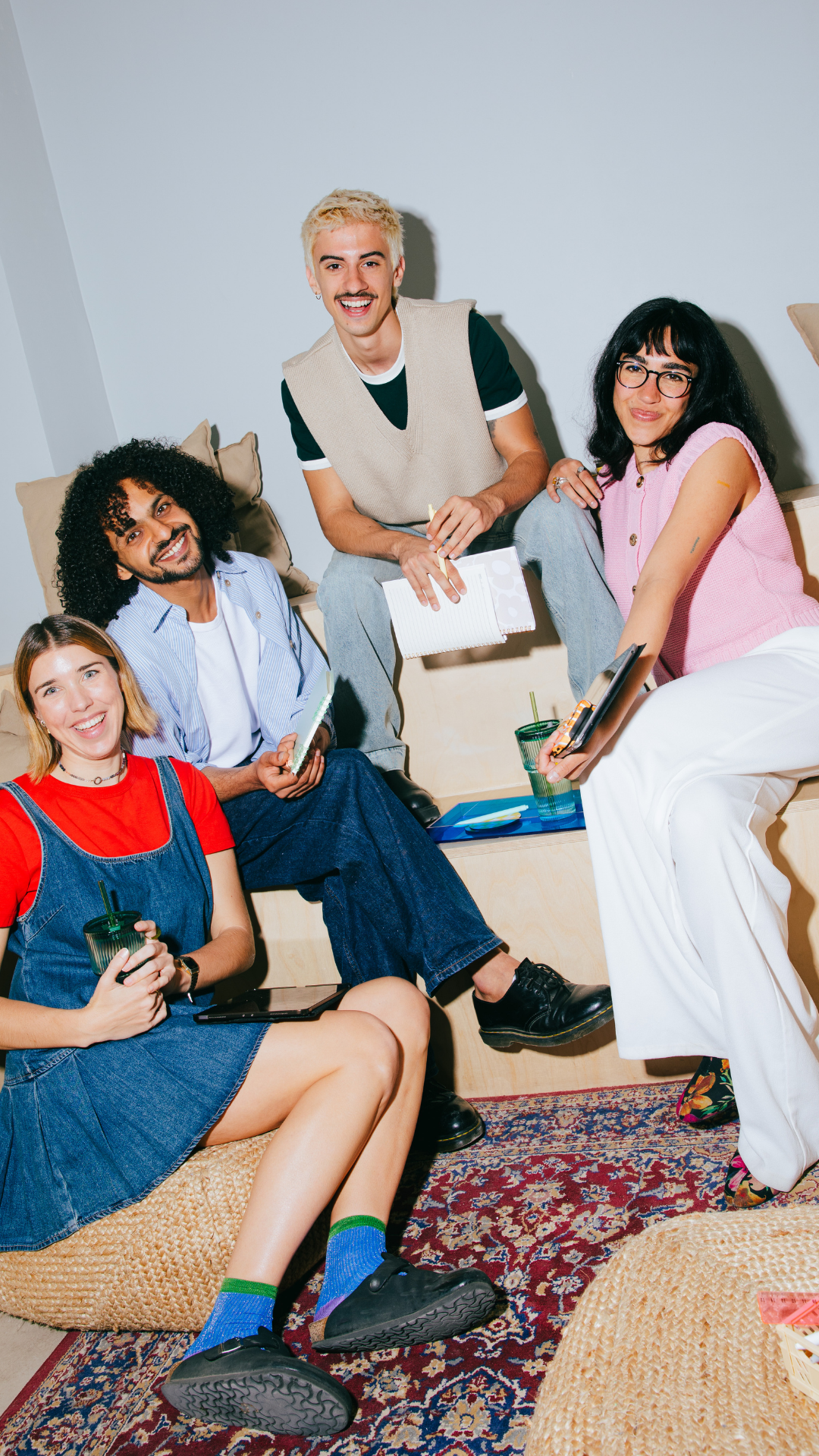 A diverse group of five young adults gathered in a cozy room, sitting and standing on and around a low wooden platform with colorful Persian-style rugs, holding drinks, notebooks, and tablets, smiling and engaging in conversation.