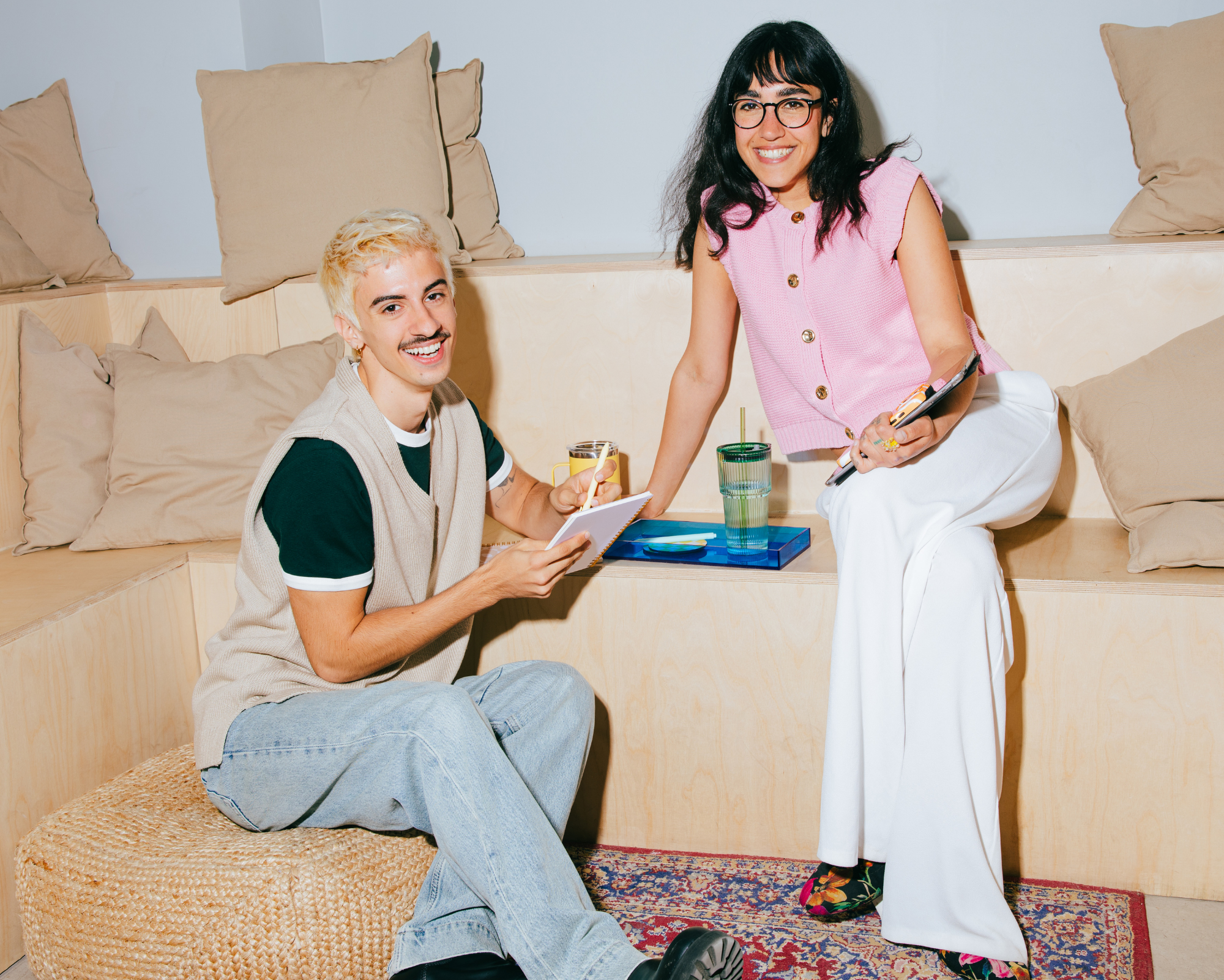 Two smiling young adults, one with short bleached blonde hair and another with long black hair, with a woman standing, holding a phone and glasses, and a man seated on a woven ottoman, holding a glass of orange juice with a straw, in a cozy room with beige pillows on a wooden bench.