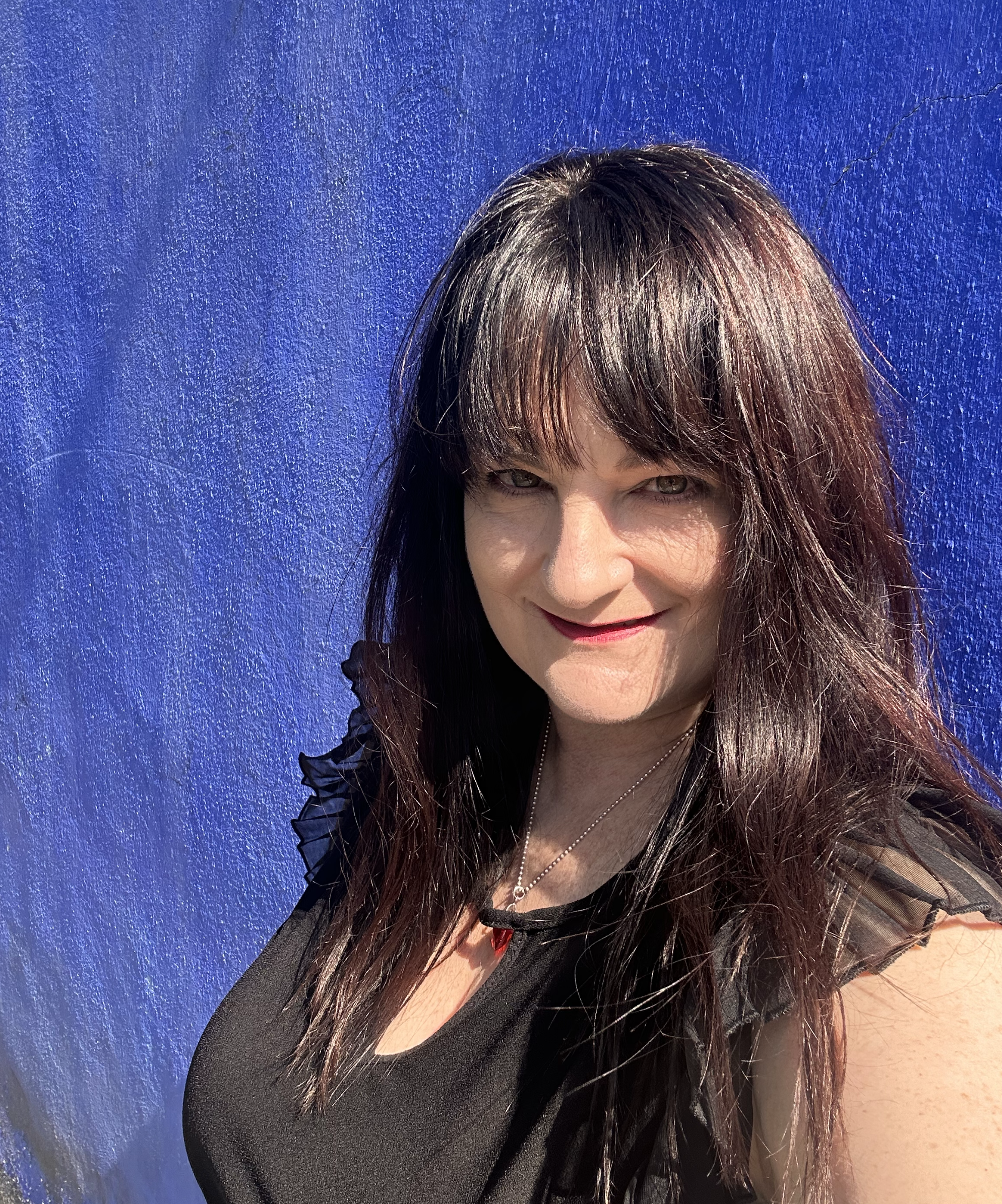 Close-up of a woman with long dark brown hair and light skin, smiling against a textured blue background, wearing a black top with ruffle details and a silver necklace.