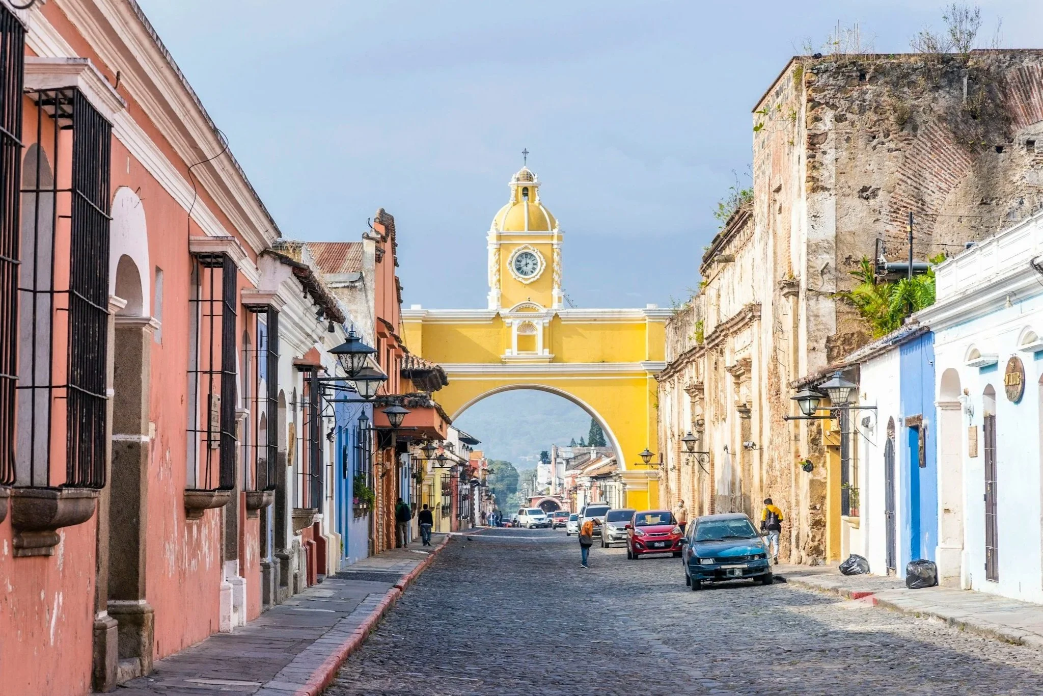 A street scene in a historic town with colorful colonial-style buildings, cobblestone road, and a yellow clock tower with a dome in the background.