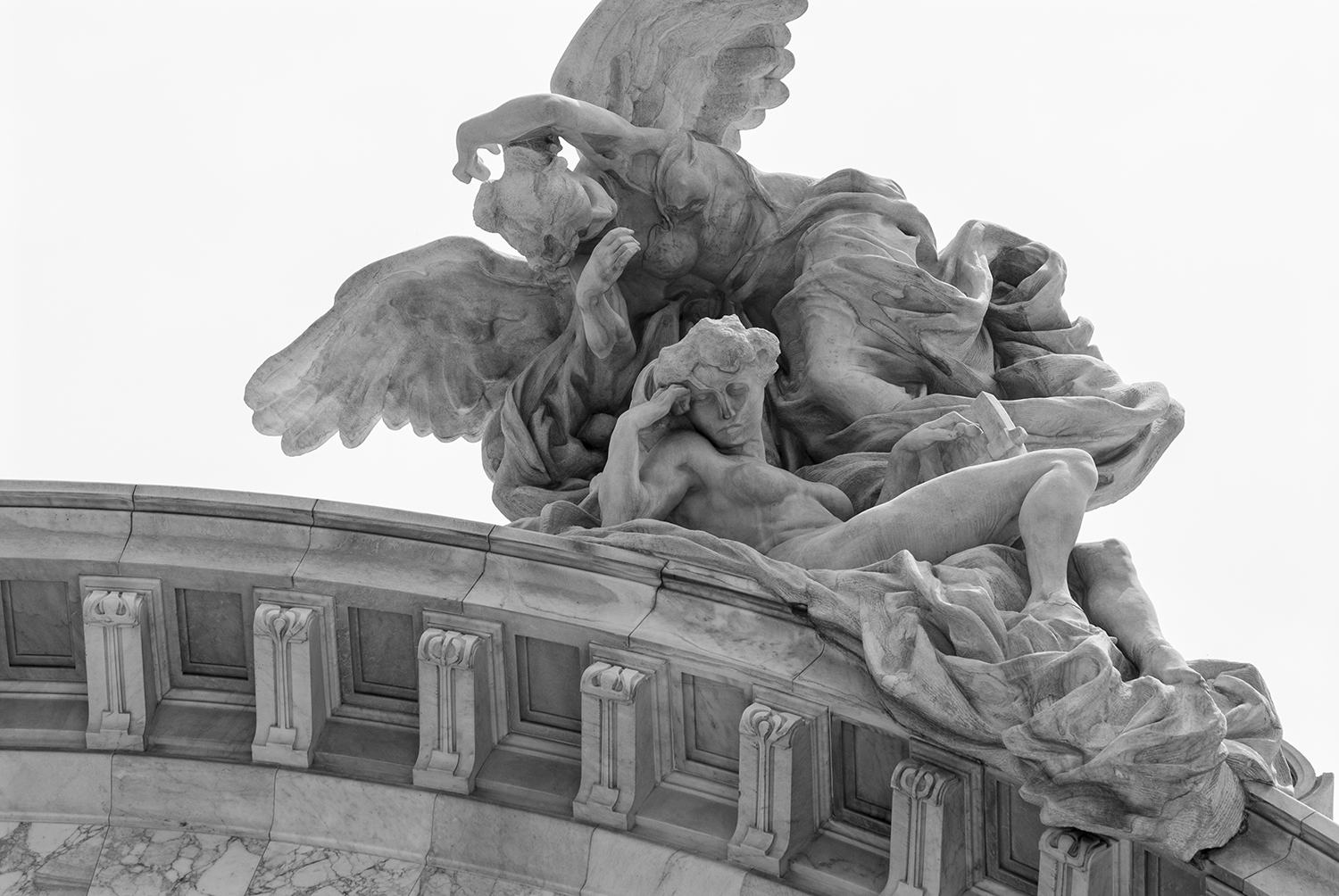 Black and white photograph of a marble statue depicting a mythological scene with a winged figure leaning over a reclining, shirtless man with a book. The sculpture features intricate drapery and detailed facial expressions, set against a plain background.
