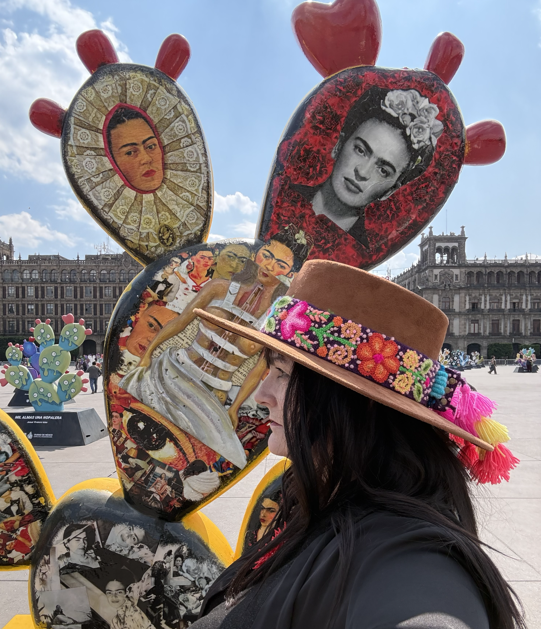 Close-up of a woman with long dark hair wearing a sombrero decorated with colorful images of Frida Kahlo, in a large plaza with historic buildings in the background.
