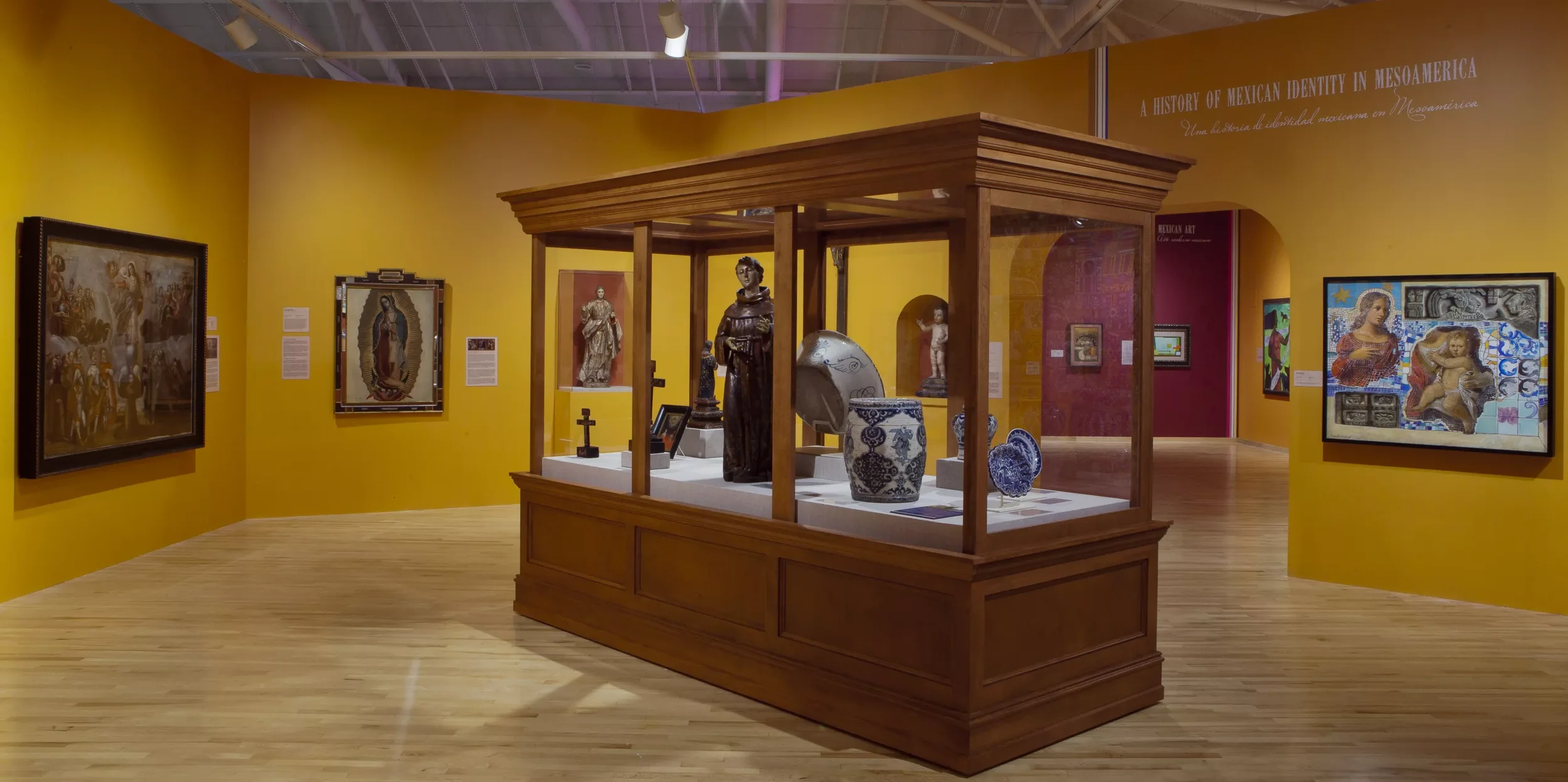 An art museum exhibit featuring sculptures, ceramics, and paintings on yellow walls. A wooden display case contains various artifacts, with a statue of a woman and decorative pottery visible. Several framed paintings are hung on the walls.