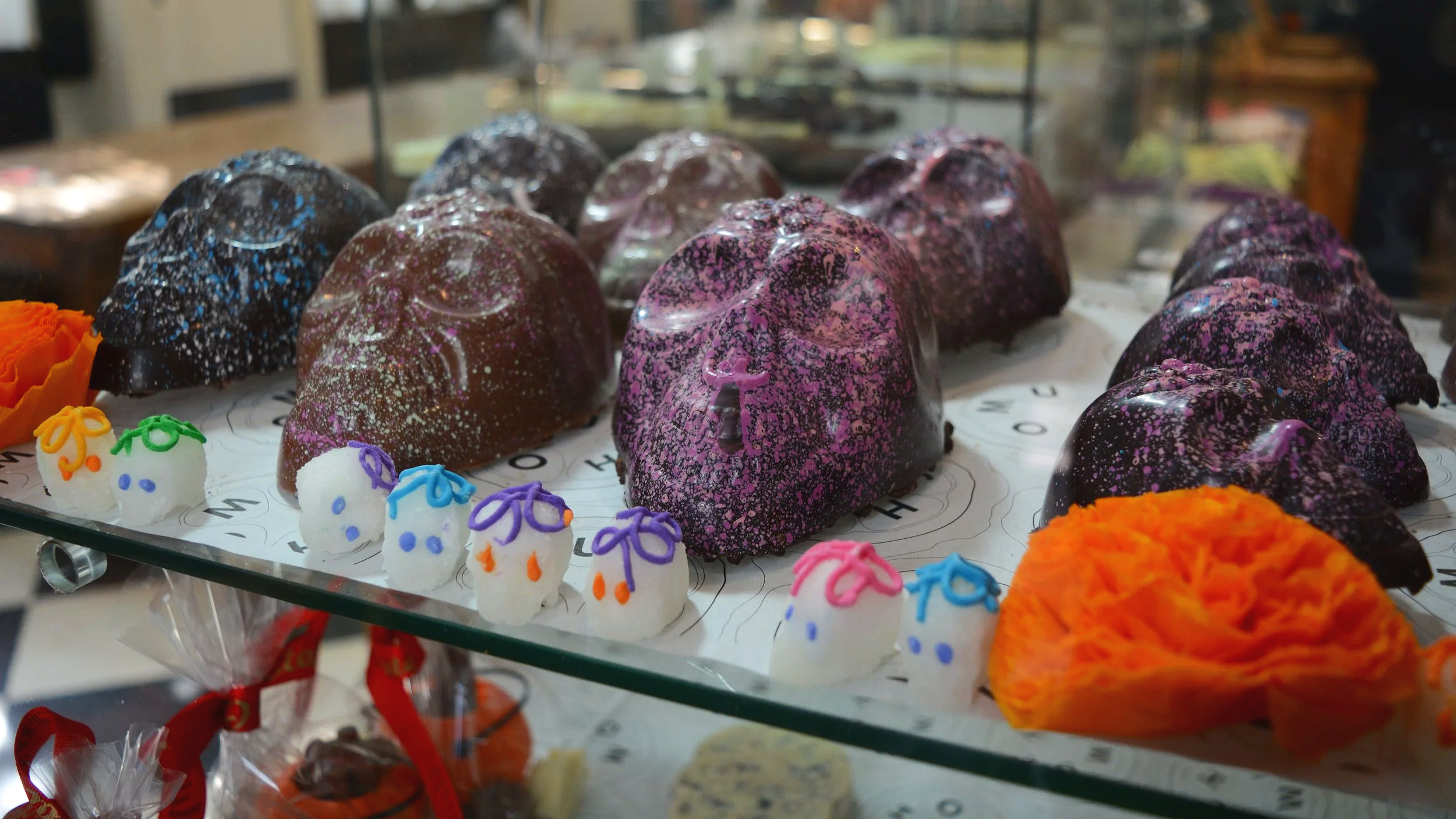 Display of Halloween-themed chocolates and candies, including ghost-shaped confections with colorful icing, decorated with purple, orange, blue, and green accents. The chocolates include shiny, speckled dark-colored pieces with a galaxy-like appearance, arranged on a glass shelf.