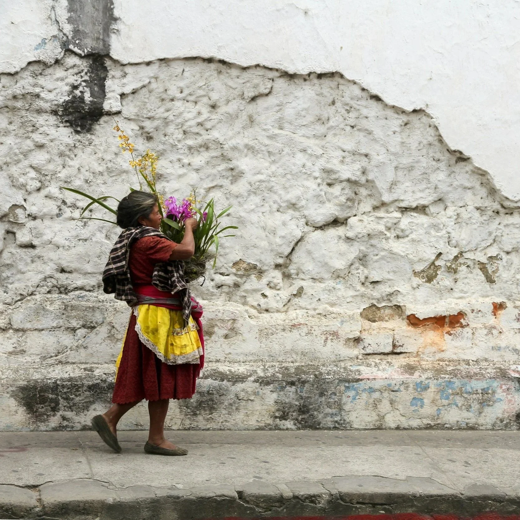 A woman with gray hair, wearing a colorful skirt and a red shirt, is walking along a textured, worn white wall while holding a bouquet of pink and yellow flowers.