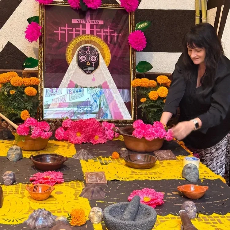 A woman arranging pink flowers on a table decorated with yellow and black cloth, in front of a large framed Dia de los Muertos artwork of a decorated skull with a dress and halo. The table has marigold flowers, small skull sculptures, and other decorative items, with colorful paper decorations hanging above.