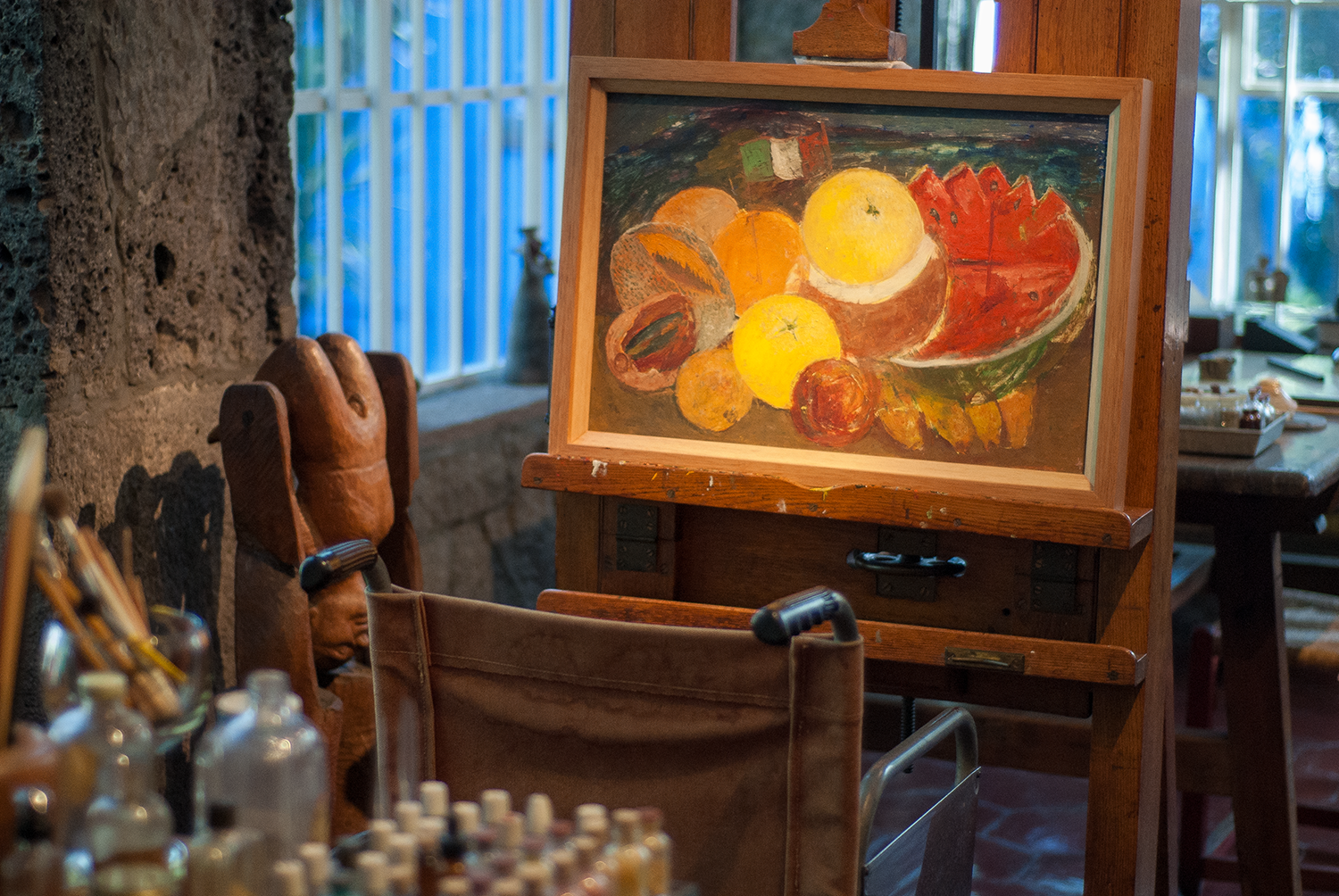 A painting of various fruits, including watermelon, lemons, oranges, and peaches, displayed on an easel in a rustic room with stone walls and large windows.