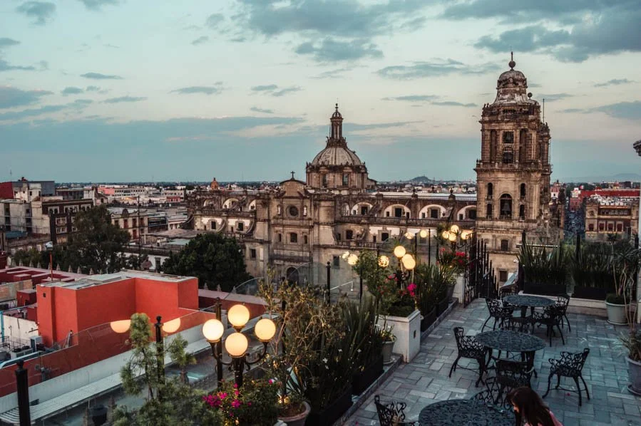 View of a historic cityscape featuring an ornate cathedral with domes and bell towers, seen from an outdoor rooftop terrace with tables, chairs, and decorative lighting.