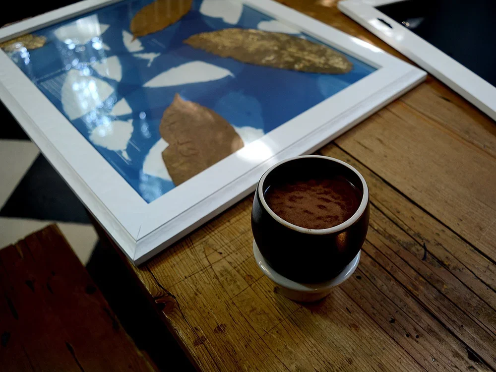 A wooden table with a framed picture of leaves, a black mug of coffee, and a tablet.