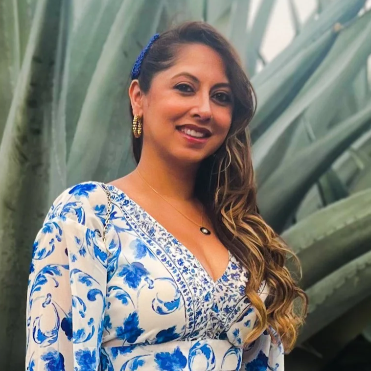 A woman with long wavy hair wearing a white dress with blue floral patterns, gold earrings, a blue headband, and a black pendant necklace, standing in front of large green plants.