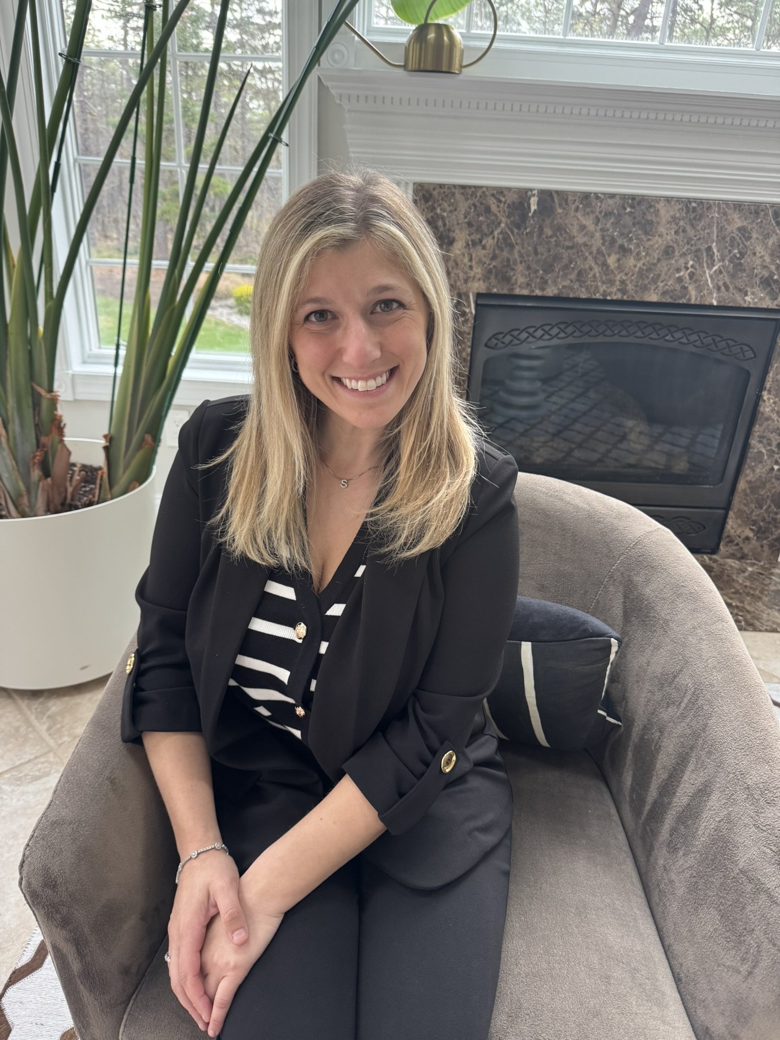 A woman with blonde hair smiling while sitting on a gray armchair in front of a window, with a large potted plant nearby and a fire place behind her.