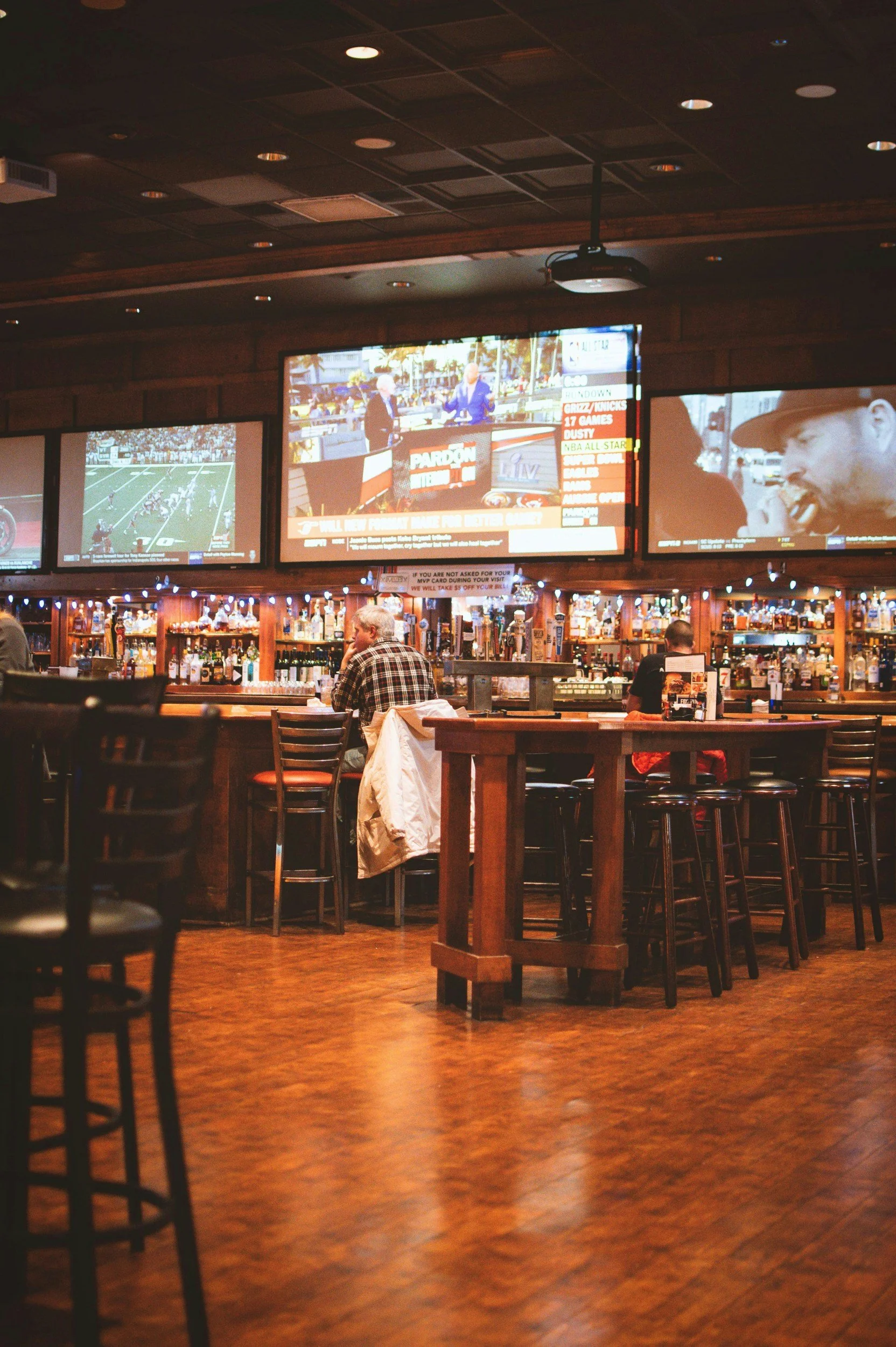 Interior of a sports bar with wooden furniture, multiple large screens showing sports and news, and patrons seated at the bar.