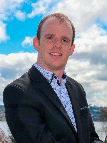 Portrait of a man in a black blazer and a patterned dress shirt standing outdoors by a body of water under a cloudy sky.