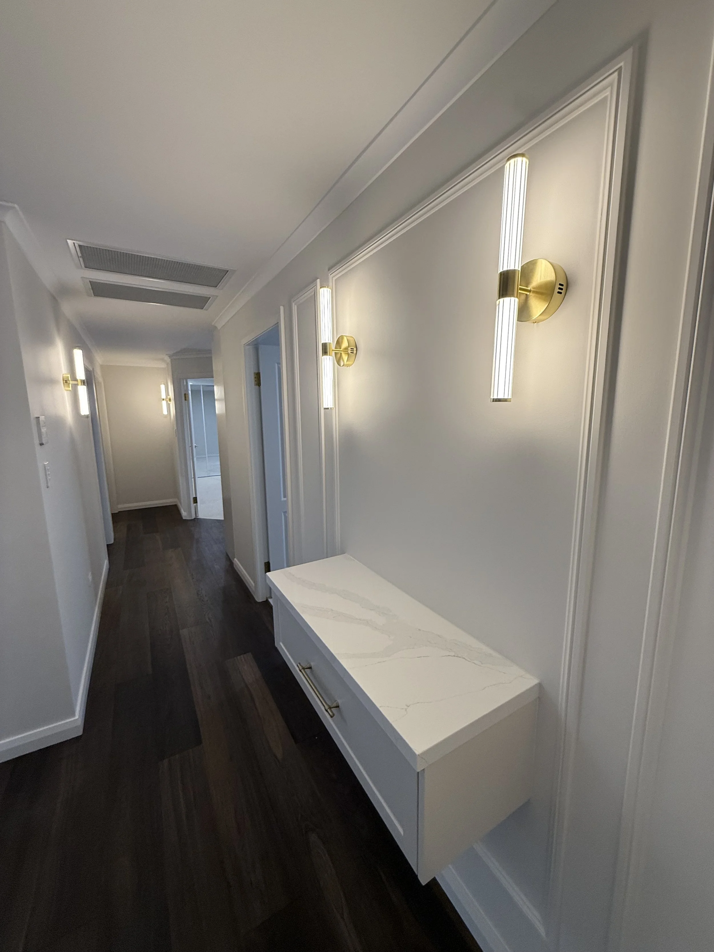 A hallway with white walls, dark wooden floor, white door frames, and modern wall-mounted gold and white lights. There's a white marble-top console table against the wall.