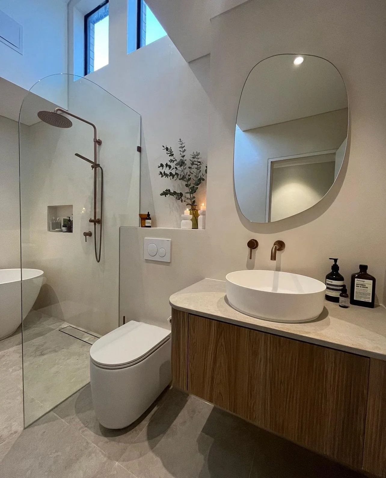 Modern bathroom with a walk-in shower, a white toilet, and a wooden vanity with a vessel sink, oval mirror, and decor including candles and bottles.