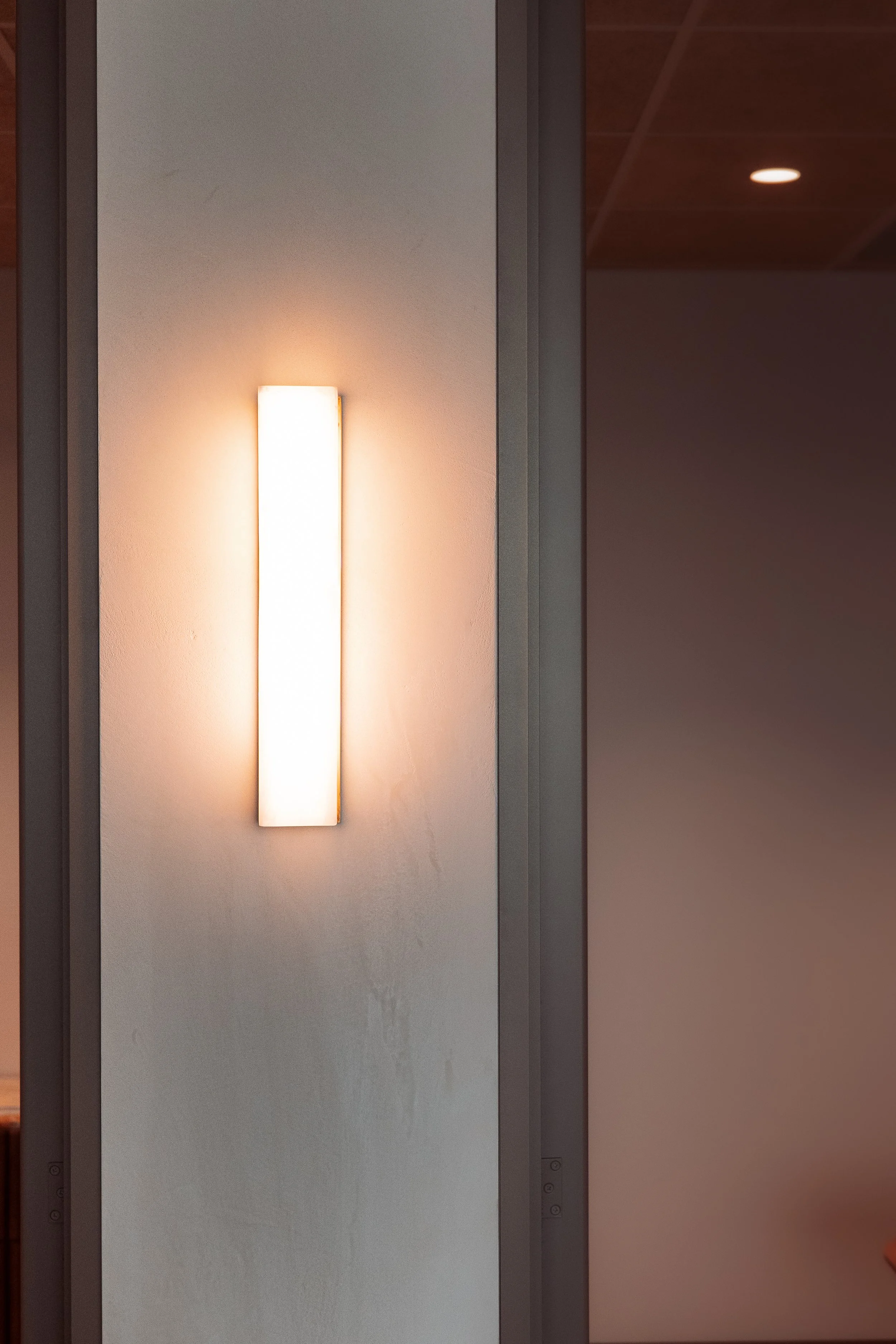 Indoor wall with a vertical rectangular light fixture emitting a warm glow, adjacent to a doorway or partition with a visible hinge and corner.