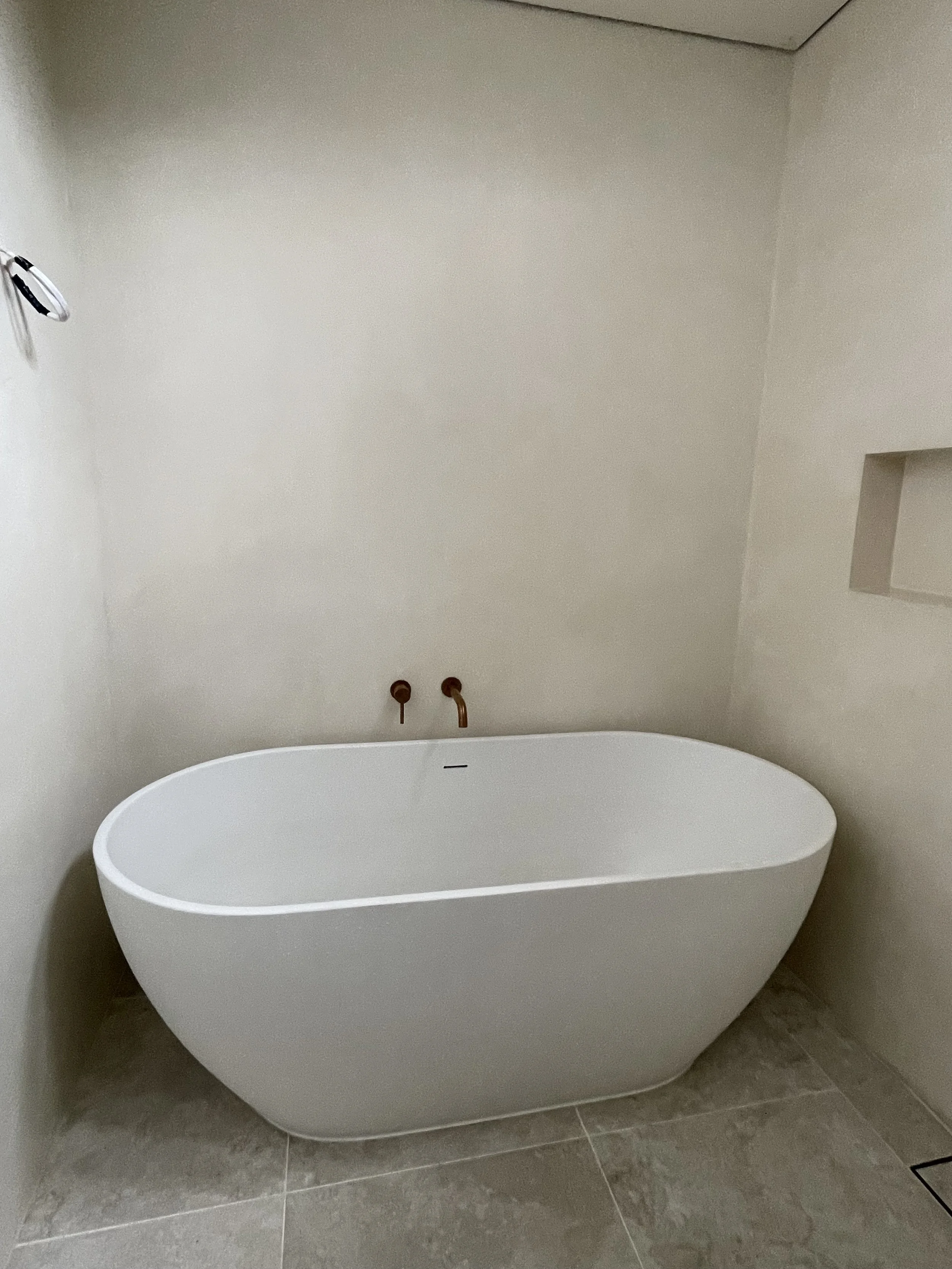 A white, modern freestanding bathtub in a bathroom with gray tiled flooring and a neutral wall. The plumbing fixtures are not visible, but there is a small rectangular niche in the wall to the right.